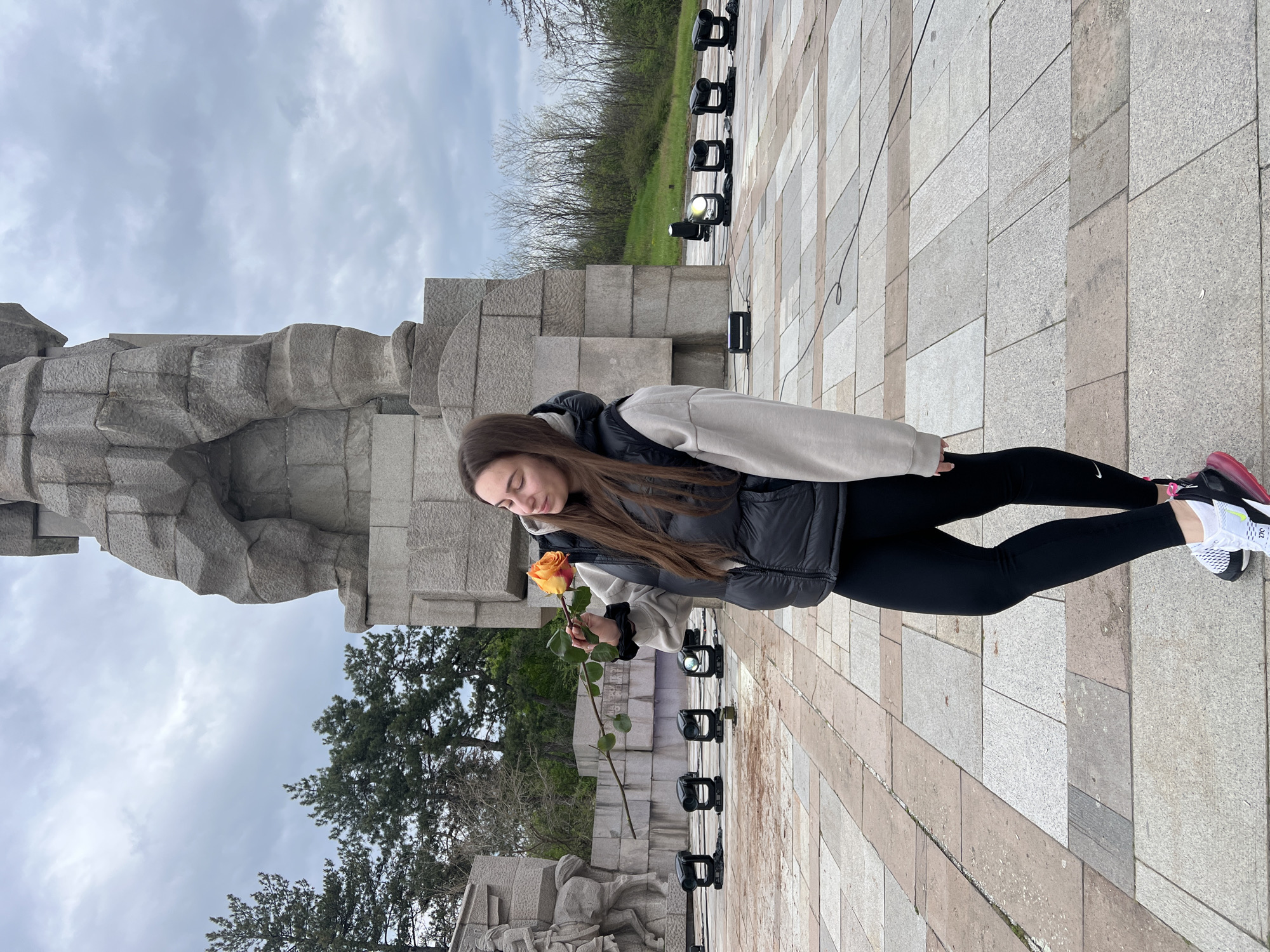 The image, taken in Panagyurishte, Bulgaria, depicts a young woman standing outdoors, holding a single orange and yellow rose. She has long, dark brown hair and is dressed in a light-colored hooded sweatshirt, a dark puffy vest, black leggings, and athletic shoes with white, black, and pink accents. Her gaze is directed gently downwards towards the rose, suggesting a moment of contemplation or peace.
To her left, a substantial grey stone monument with a rugged, blocky, and somewhat abstract design forms a prominent backdrop, likely a memorial or historical structure. Below the main structure, carved figures in relief are visible. The ground is paved with square and rectangular tiles in shades of grey and beige. To her right, a line of modern black outdoor lighting fixtures is arranged on the tiled surface, with one light emitting a bright beam. A black cable runs along the ground. In the distance, beyond the monument, an area with bare trees and some green foliage can be seen under an overcast sky, suggesting a cool, cloudy day. No significant text is clearly visible.