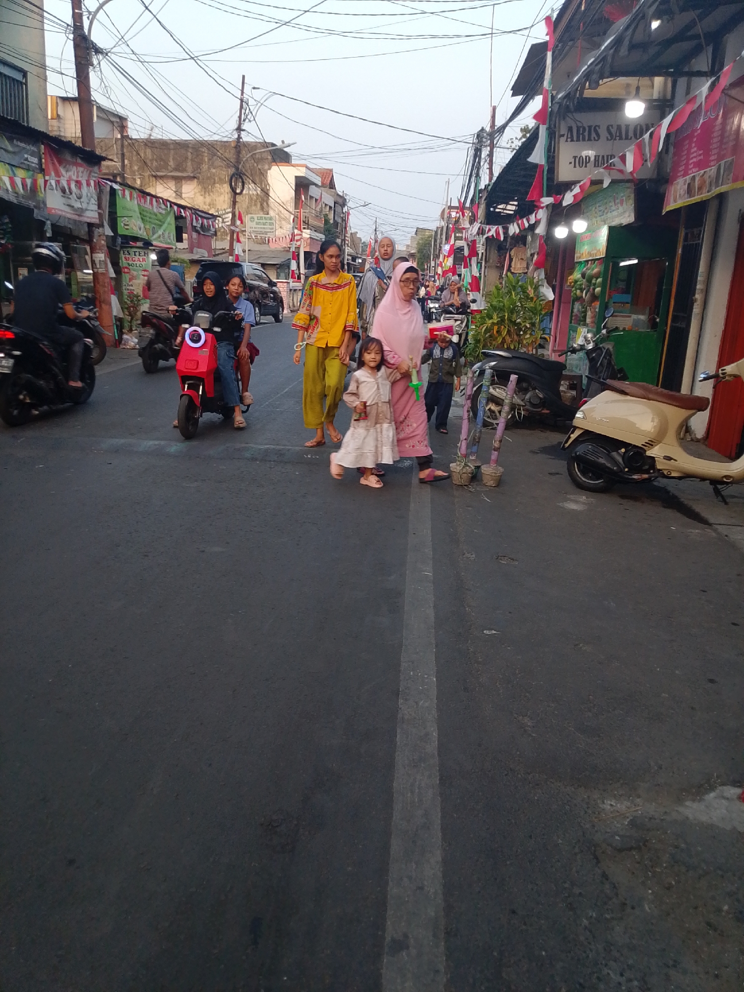 In East Jakarta, Indonesia, a street scene unfolds as several people walk on a paved road lined with shops. In the foreground, a young girl in a light-colored dress walks alongside an adult woman wearing a pink headscarf and a pink outfit. To their left, another woman in a yellow shirt and green pants walks with two children. Behind them, two individuals ride a red electric scooter. Further down the street, a person rides a black motorcycle, and a beige scooter is parked on the right side of the road. The shops lining the street display various signs and decorations, including Indonesian flags. The sky above is overcast.