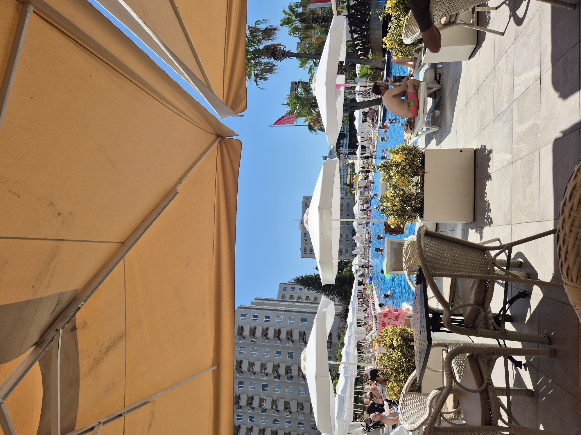 The image depicts a crowded outdoor swimming pool area on a sunny day. Multiple white umbrellas and large tan awnings provide shade over lounge chairs and seating areas. In the background, a multi-story building with many balconies is visible, suggesting a hotel or resort. Palm trees are scattered throughout the scene.
The pool is filled with people swimming, lounging, and interacting. Some individuals are seen sunbathing on lounge chairs around the pool. In the foreground, there is a table with several chairs made of woven material. The ground is tiled.
The overall scene suggests a leisure environment, likely a resort, where people are enjoying a vacation or recreational activities. The presence of numerous guests and amenities points to a busy and vibrant atmosphere.
