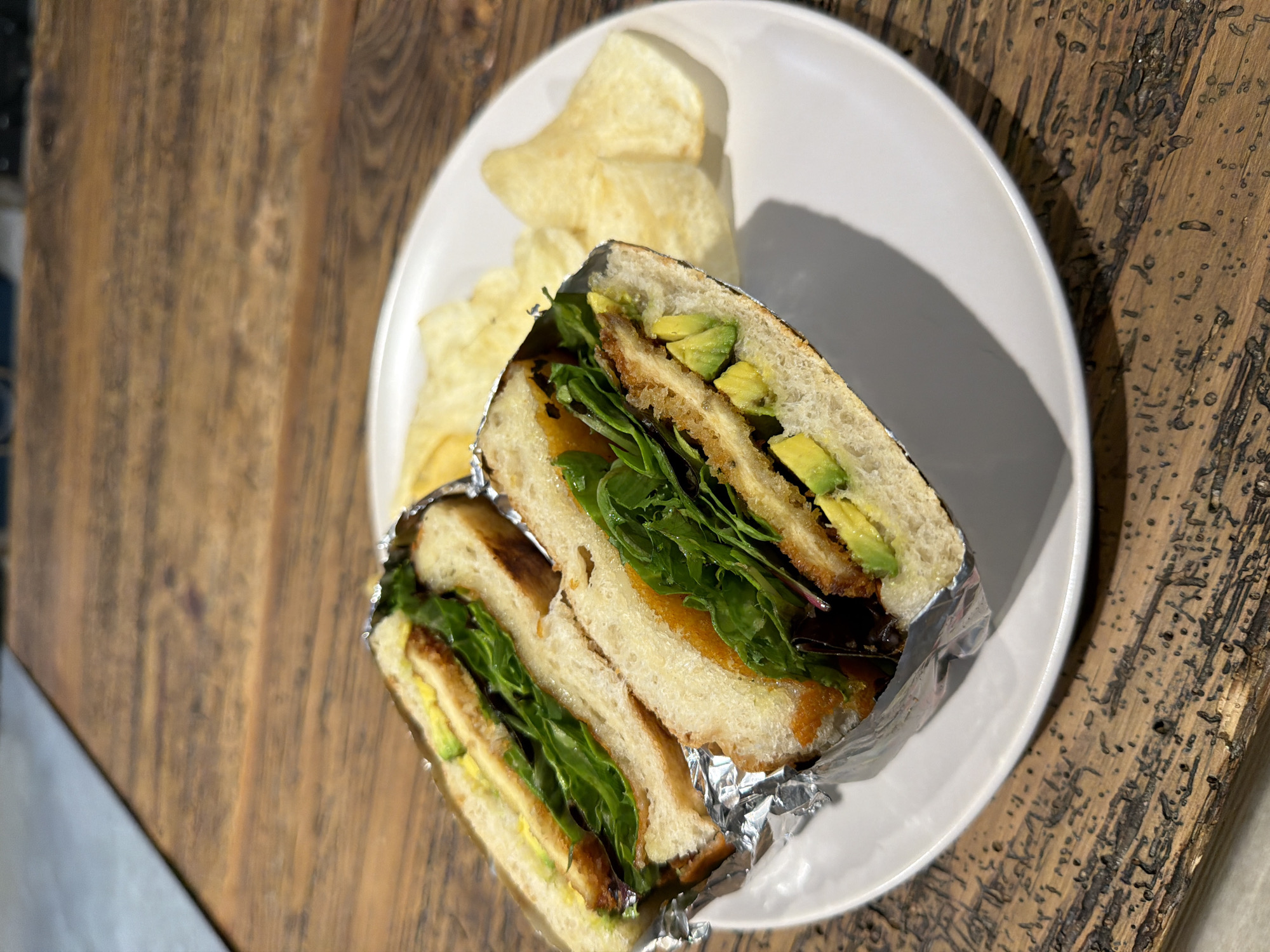 A halved sandwich, wrapped in foil, sits on a white plate alongside a small pile of potato chips. The sandwich appears to contain breaded meat, avocado slices, and greens. The plate is placed on a weathered wooden surface. The scene is illuminated by a light source, casting a distinct shadow behind the plate. The setting may be a restaurant in Puebla, Mexico, but there is no definitive evidence in the image to confirm the location.