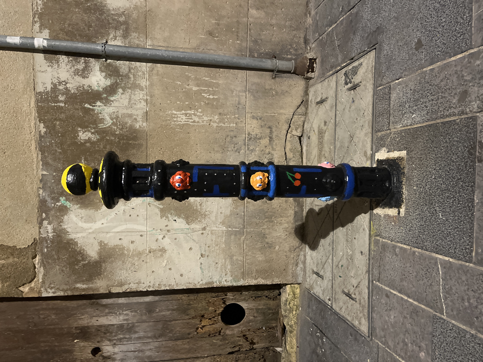 A black bollard, decorated with blue and white painted patterns and various cartoonish characters in red, orange, and pink, stands on a sidewalk in Tarragona, Spain. The characters appear to be inspired by Pac-Man ghosts and cherries. The bollard is topped with a black sphere adorned with a yellow and black pattern resembling a simplified Pac-Man. The bollard casts a shadow to its left on the sidewalk, which is paved with square and rectangular tiles. Behind the bollard is a textured, light-colored wall, partially weathered and stained. To the left of the bollard, a dark wooden door with a circular opening is visible. To the right, a gray metal pipe runs vertically along the wall, attached by metal clamps. The scene appears to be an urban street at night or in low light conditions.