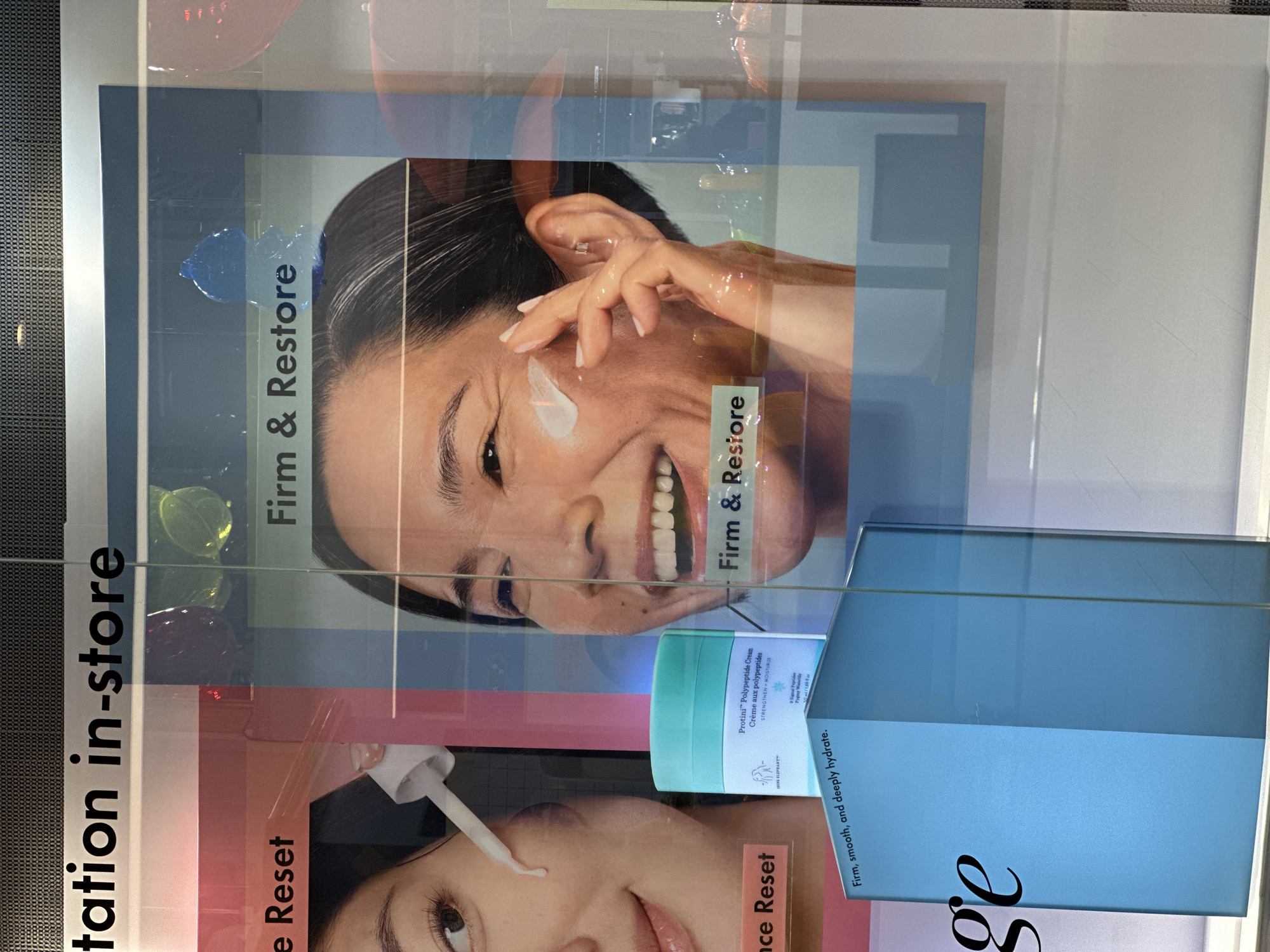 The image shows a close-up of an advertisement display, likely inside a store. The advertisement features beauty products, specifically creams, designed to firm, restore, and hydrate the skin. The products are displayed alongside images of women using the creams. The display is likely located in Bangkok, Thailand, though this is not directly visible in the image. The products appear to be skincare creams from the brand "Drunk Elephant," as evidenced by the branding on the packaging. The environment appears to be a store window or display case, with reflections and other objects visible in the background.