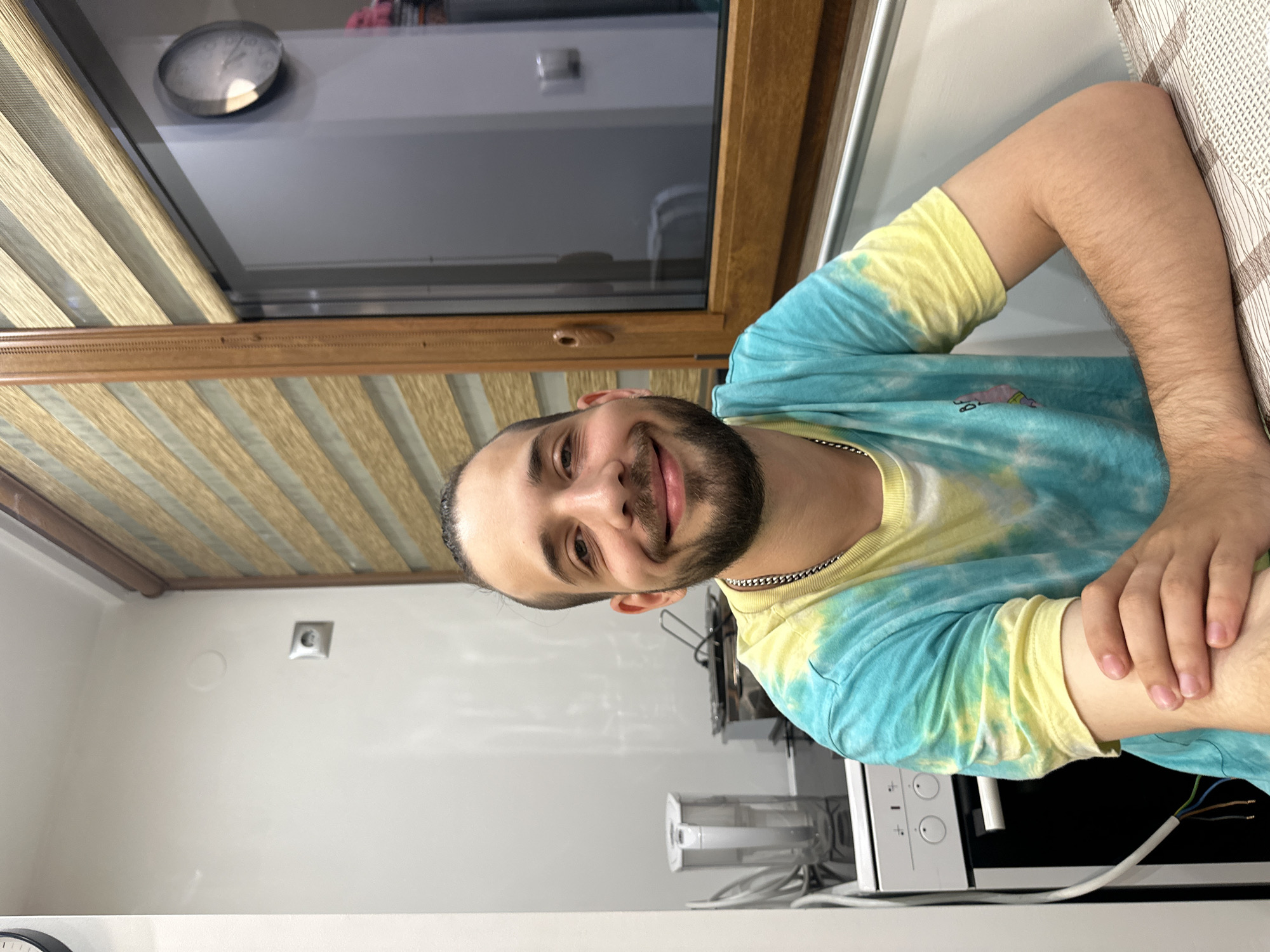 A man with short dark hair and a beard is positioned to the right and in the foreground of the image, smiling directly at the camera. He is wearing a tie-dye shirt of blue and yellow. His arm is resting on a surface, possibly a counter or table, and his hand is visible.
Behind the man, a window is visible with a light-colored blind partially obscuring the view. The window frame is brown. A clock is affixed to the wall above the window. Below the man and window, a white wall is visible. Additionally, there is a white microwave to the right, and a white kitchen appliance with a transparent container in the background, along with some cables.
The image does not allow for determining the location in the city of Lovech, Bulgaria.