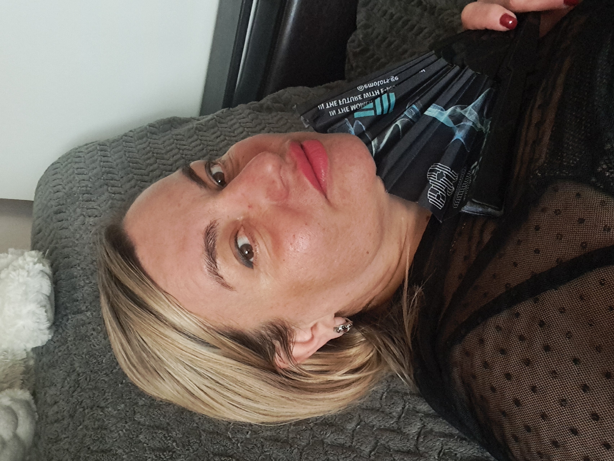 A woman with blonde hair lies on a gray textured surface, possibly a bed or couch. She wears a black sheer top. A folded, black and blue patterned object, possibly a fan or a stack of papers, rests against her neck and shoulder. A portion of a white object is visible in the lower left corner of the image. No specific location in Elektrostal, Russia can be determined from this image.