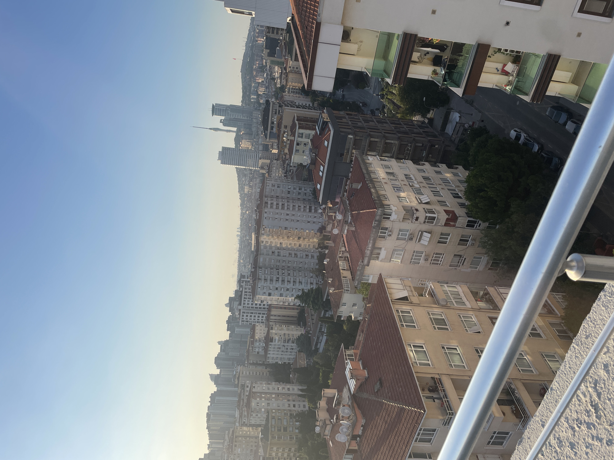 This image captures a panoramic view of Kadıköy, Turkey, likely taken from a high vantage point during the late afternoon or early evening. The city skyline is dominated by a dense collection of apartment buildings of varying heights and architectural styles. Many buildings feature red-tiled roofs, a common characteristic of Turkish urban landscapes. Some buildings have balconies, and a few are adorned with satellite dishes. In the distance, modern skyscrapers and a prominent telecommunications tower stand out, suggesting a blend of older and newer urban development. The sky is a clear, pale blue, transitioning to a warmer tone near the horizon, indicating the setting sun. A few cars are visible on a street winding through the buildings. The foreground is partially occupied by a modern balcony railing, made of polished metal, and a textured concrete surface, hinting at a residential setting. No individuals are clearly discernible in the image. The overall impression is one of a bustling urban environment bathed in soft, natural light.