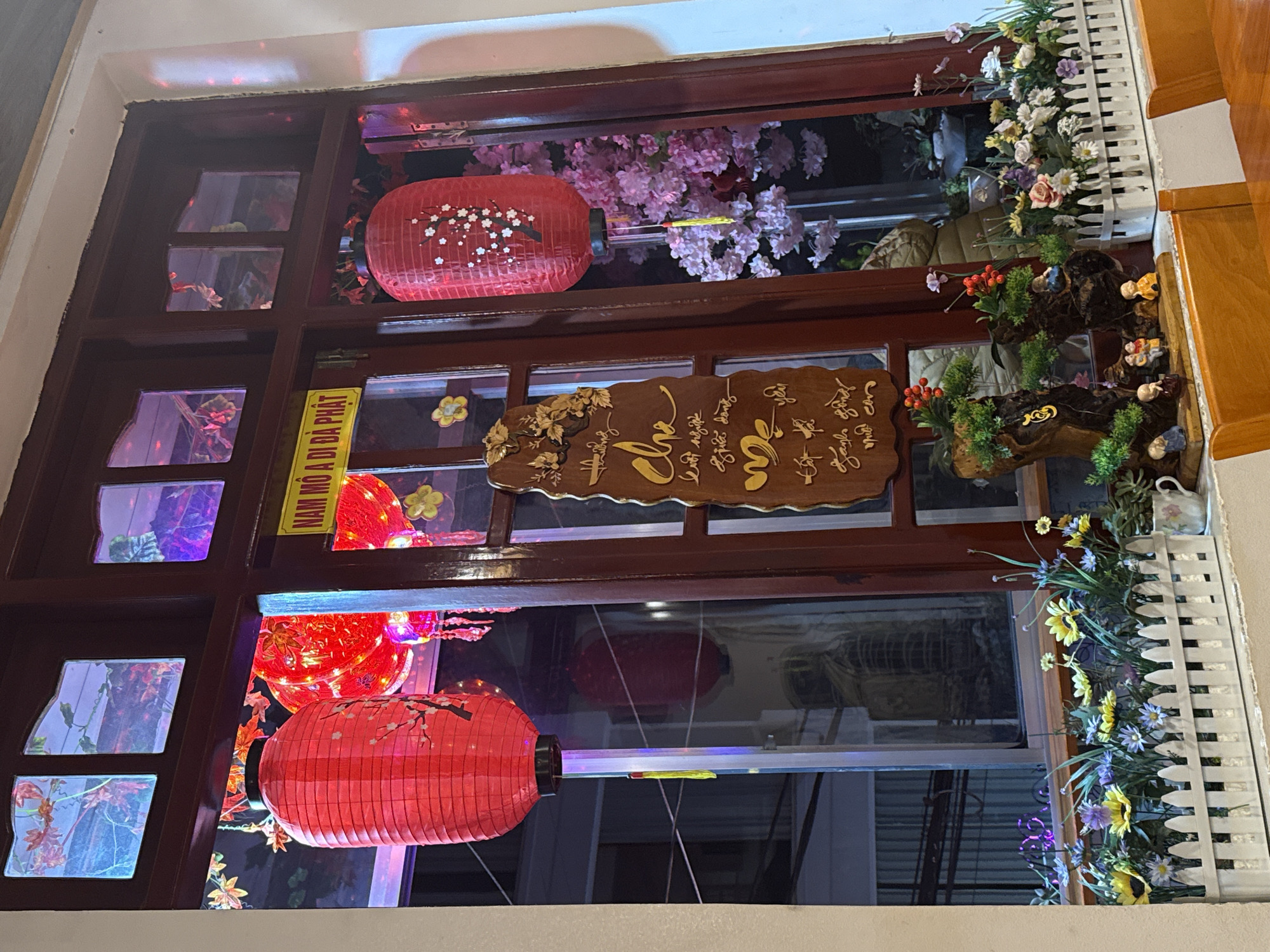 This detailed image depicts a decorated wooden-framed window located in Da Lat, Vietnam, viewed from an interior perspective. The window frame is dark brown wood, divided into multiple panes. The upper panes feature translucent, textured glass with leaf patterns, illuminated with blue and purple light.
Through the main window panes, several red paper lanterns are prominently displayed. On the left, a large cylindrical red lantern adorned with white cherry blossom motifs and a yellow tassel hangs. To its right, another similar lantern is visible, partially obscured. Other smaller, glowing red lanterns are also present behind the larger ones.
Affixed to the central window frame is a rectangular yellow sign with black Vietnamese text that reads "NAM MÔ A DI ĐÀ PHẬT" (Namo Amitabha Buddha). Below this sign, a vertical, irregularly shaped wooden plaque with intricate gilded Vietnamese calligraphy is mounted.
The window sill is adorned with an elaborate display. On the left, a small collection features miniature bonsai-like trees with red berries, and several small figurines, including robed figures resembling monks and a group of three individuals. A white ceramic mug is placed beside this display. To the right, a white planter with a miniature picket fence holds an arrangement of artificial flowers, including yellow, purple, and white daisies. Visible through the right window pane are artificial pink cherry blossom branches.
Reflections on the window glass show parts of the interior space, including a wooden table and chair in the foreground. Through the clearer glass sections, a glimpse of another building or structure with windows is visible in the background, suggesting an urban or residential setting. No people are visible in the scene.