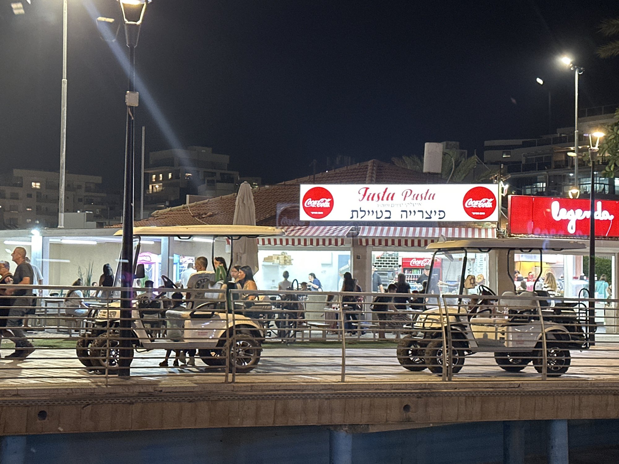 This image depicts a night scene at an outdoor promenade in Nahariyya, Israel. In the foreground, two golf carts are parked on a wooden boardwalk. The cart on the left has a woman in the passenger seat and a man driving. The cart on the right has several individuals seated inside. Behind the golf carts, a railing separates the promenade from a seating area where several people are gathered at tables.
Beyond the seating area, a building features a sign for "Fasta Pasta" with Coca-Cola logos on either side. The sign is illuminated and appears to be for a restaurant. To the right of the "Fasta Pasta" sign is a bright red sign with "legenda" written in white script, also illuminated. In the background, several multi-story buildings are visible, with some windows lit up, indicating residential or commercial spaces. Streetlights are present, casting light on the scene. The overall atmosphere suggests a lively evening in a coastal city.