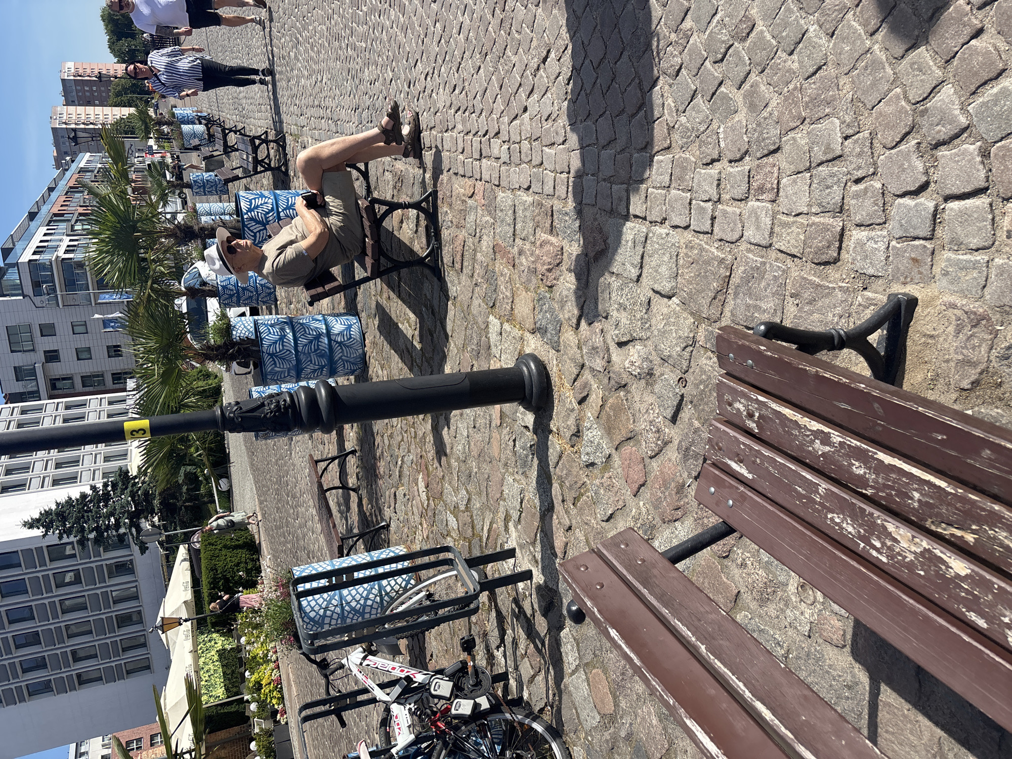 A group of people are enjoying a sunny day outdoors. In the foreground, a person is sitting on a wooden bench with their legs extended, wearing a hat and sunglasses. Beside them, another person is sitting on a bench, holding a phone. In the background, two people are walking on a cobblestone path. One person is wearing a striped shirt, and the other is wearing a white shirt and dark shorts.
The scene appears to be a public area, possibly a park or promenade, with trees and buildings visible in the distance. Several decorative planters with blue and white patterns are placed along the path. A lamppost stands near the bench in the foreground. A bicycle is parked next to one of the planters. The overall mood seems relaxed and casual, suggesting a pleasant afternoon outing. The bright sunlight and shadows indicate it is daytime, likely midday or early afternoon. There is no visible text in the image.