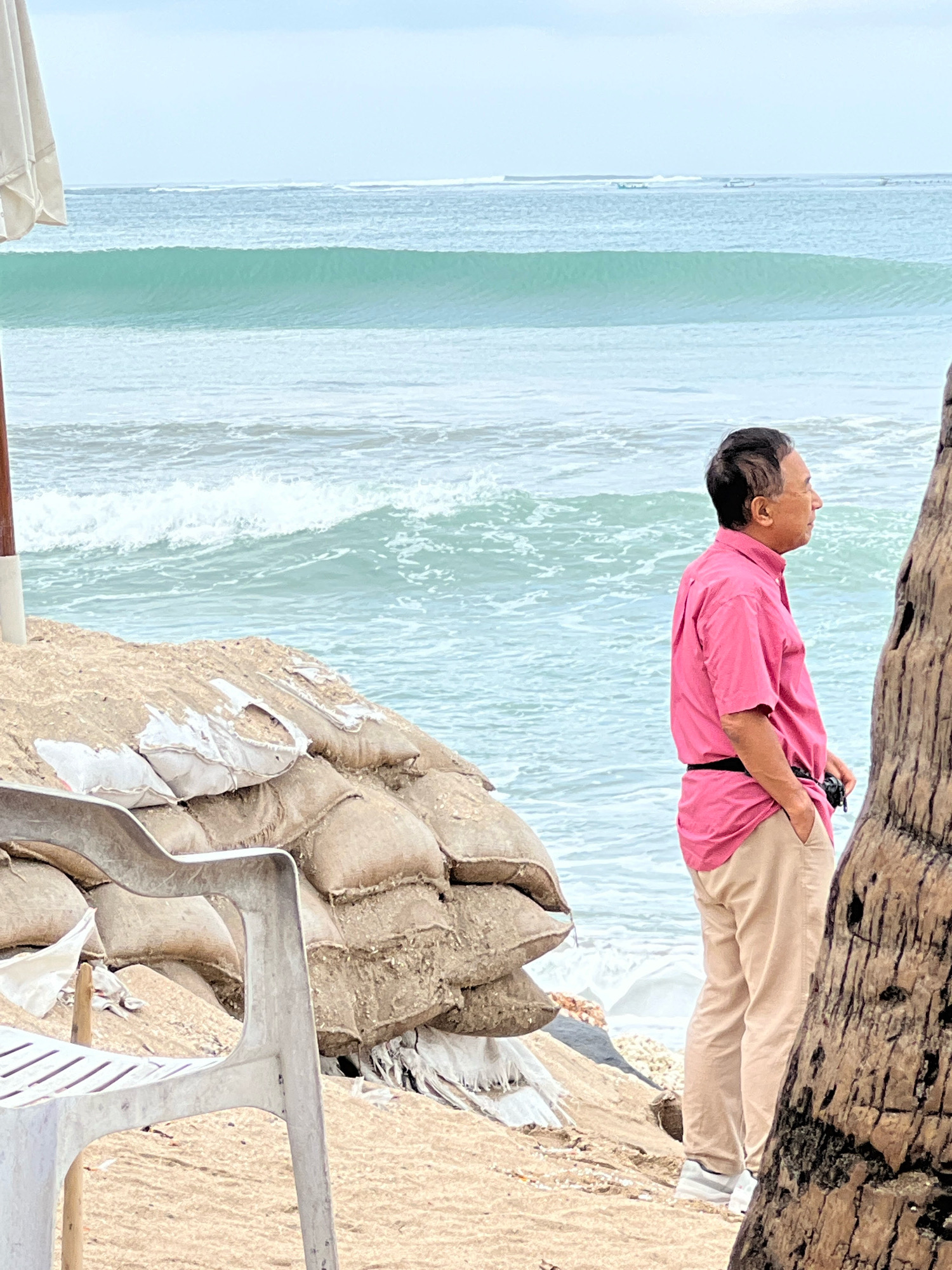 A man in a pink short-sleeved shirt and tan pants stands on a beach in Kuta, Indonesia, looking out at the ocean. Sandbags are piled up behind him, and a white plastic chair is in the foreground. The ocean has calm turquoise waves. The sky is overcast. A palm tree is on the right side of the frame.