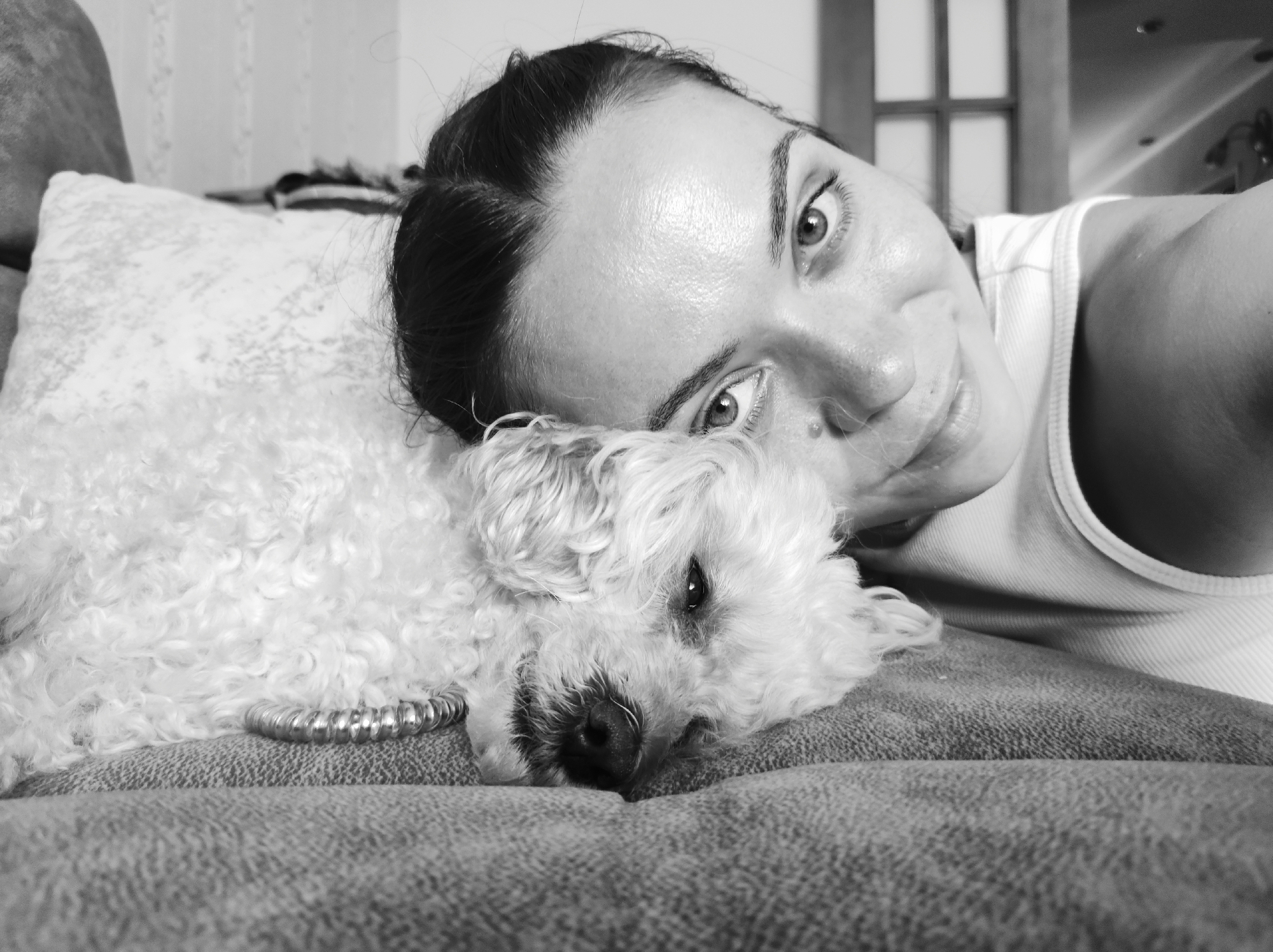 A black and white, medium close-up selfie shows a woman lying on a couch, resting her head on a small white poodle. The woman is on the right side of the frame, with her face angled towards the camera. She has dark hair pulled back and is wearing a white tank top. Her eyes are open and looking at the camera. The poodle is on the left, with its head down, appearing to be asleep or resting. A silver spiral-shaped hair tie is visible around the dog's neck. The couch appears to be made of textured fabric. In the background, a wall with a textured pattern is visible, along with a doorway or window. The image appears to be taken indoors, likely in a residential setting in Chaikovsky, Russia, given the context.