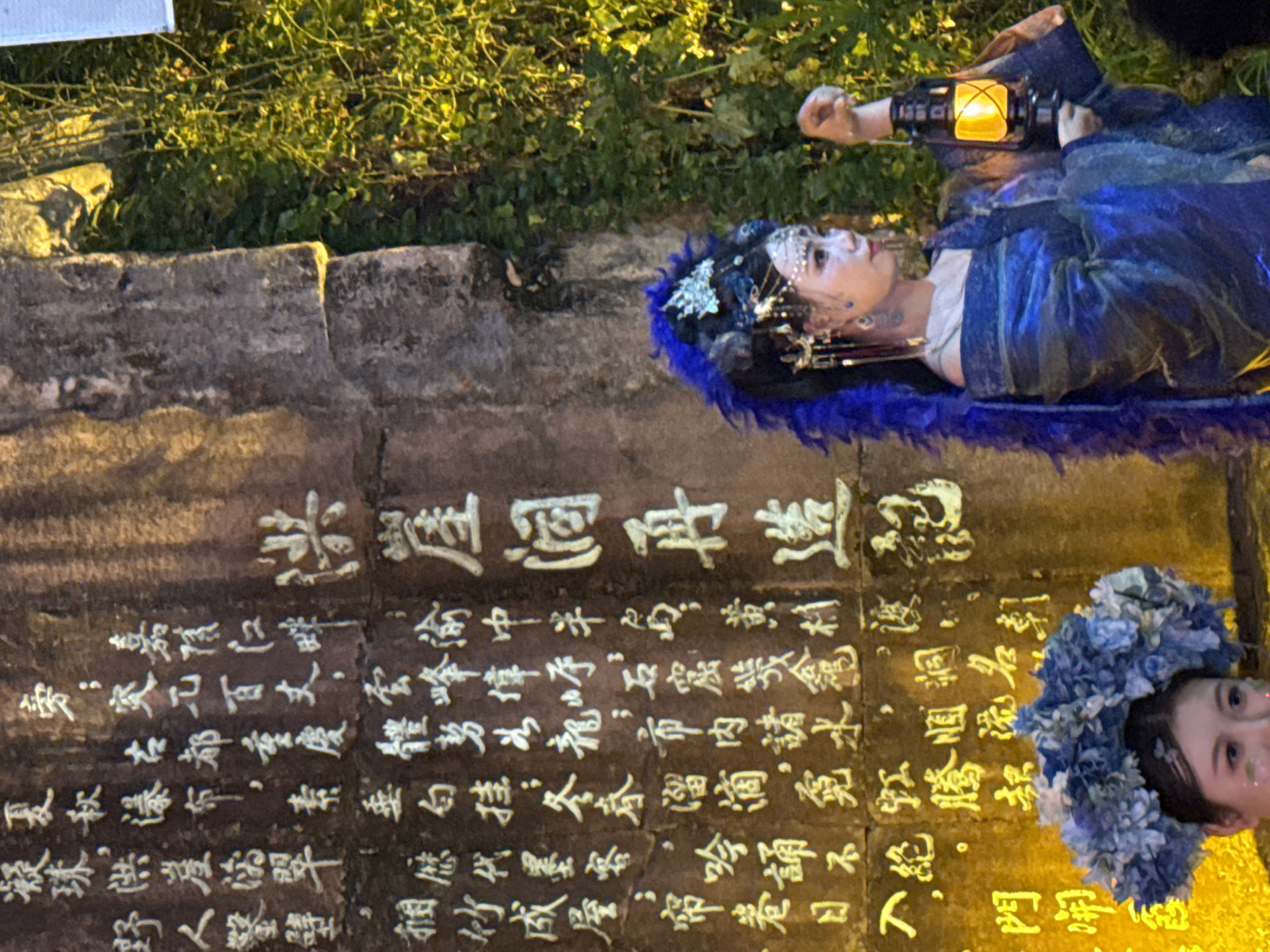 In Yuzhong District, China, a person dressed in a blue, flowing traditional outfit with elaborate headwear is holding a lit lantern. The person stands in front of a large stone monument inscribed with Chinese characters. The monument appears aged, with some of the inscriptions weathered. Behind the monument and to the right, lush green foliage is visible, suggesting an outdoor setting with natural elements. The overall lighting suggests it is either dusk or nighttime, with artificial light from the lantern and potentially other sources illuminating the scene.