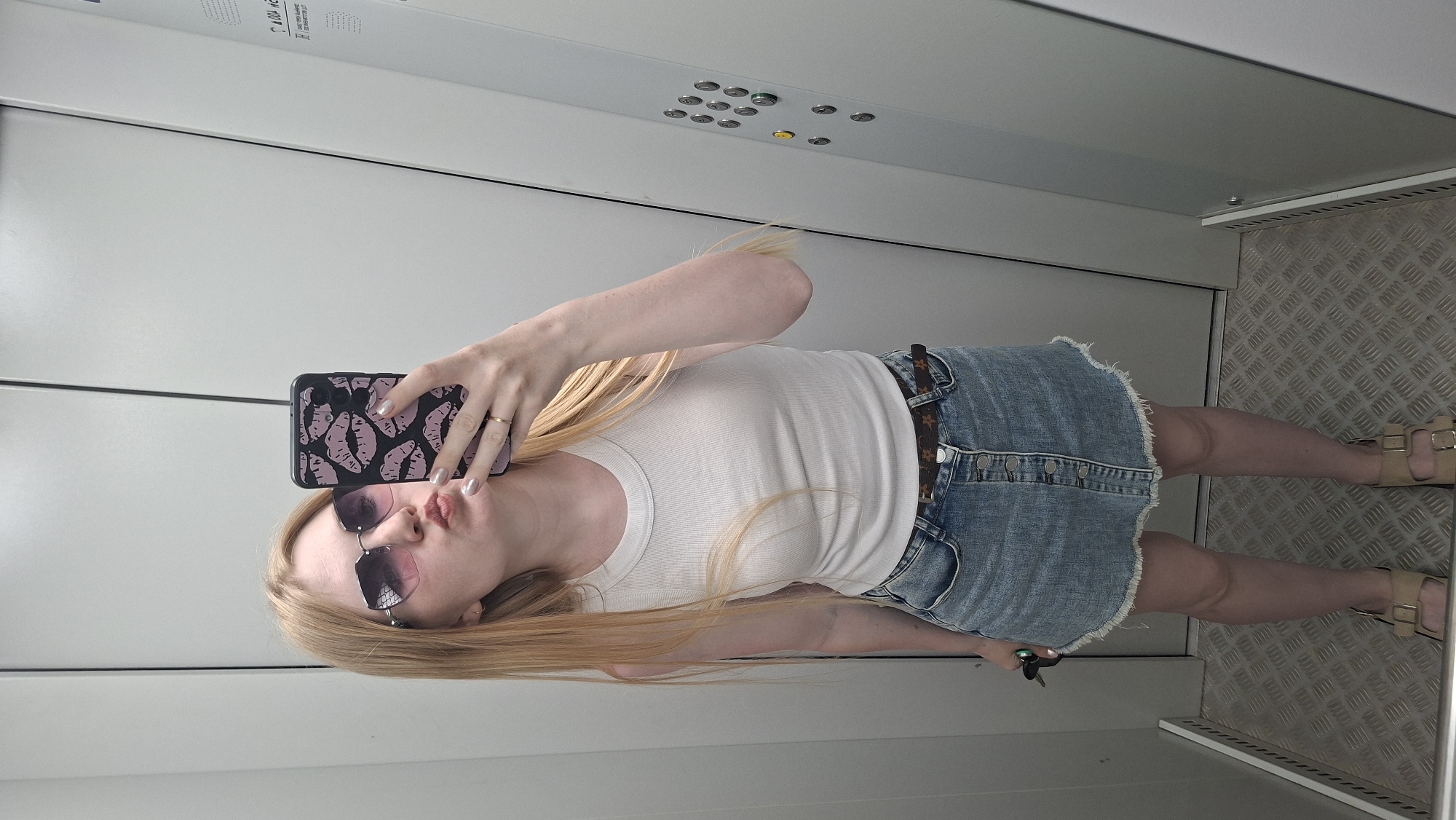A full shot depicts a female in an elevator. The female is fair-skinned and blonde, wears tinted sunglasses, a white tank top, a blue jean miniskirt, tan sandals and holds up a cellphone with a lip pattern case. The elevator has white walls and diamond-patterned steel floor. Elevator buttons are visible on the wall.