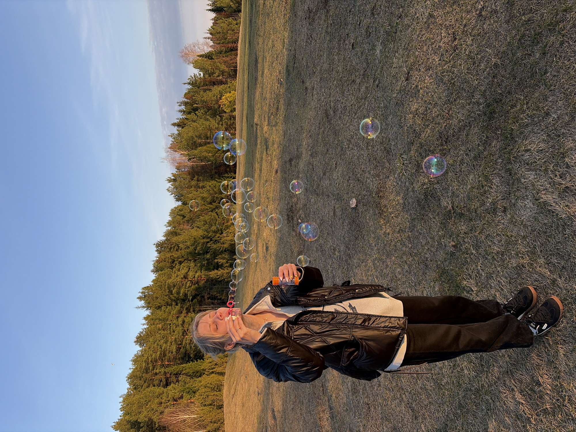 A person is blowing bubbles outdoors in a park or field in Ольховка, Russia, during the daytime. The subject is wearing a black jacket, light grey sweater, black pants, and black sneakers with brown soles. They are holding a small orange bottle, presumably containing bubble solution, and blowing bubbles with a wand. Numerous iridescent bubbles are visible floating in the air. The background shows a grassy field with a line of trees in the distance, and a blue sky with light clouds is visible. The image has an aspect that suggests it was taken at or around sunset as the sun seems to be low on the horizon.