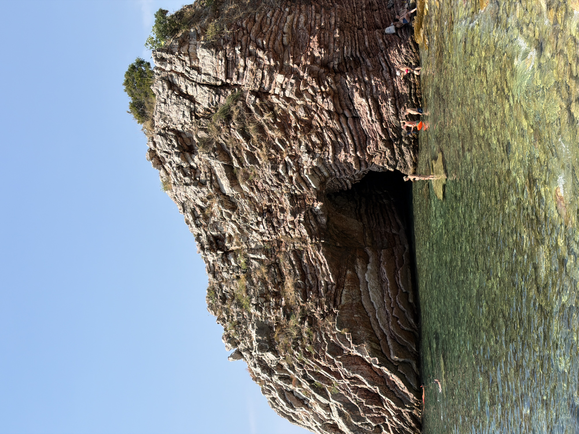 The image shows a large, layered rock formation with a cave opening, jutting out into clear, shallow water. The rock is reddish-brown and has many horizontal striations. Vegetation, including small trees and dry grass, grows on top and in crevices of the rock. Several people are visible in and around the water. In the water, two people are swimming or wading, and a red inflatable object is also present. Near the cave entrance, a group of people are standing on rocks or in shallow water, some appearing to be interacting. Further to the right, two individuals are seated on a rock. The sky is clear and blue. The scene is located at the entrance to a sea cave at the coast of Boreti, Montenegro.