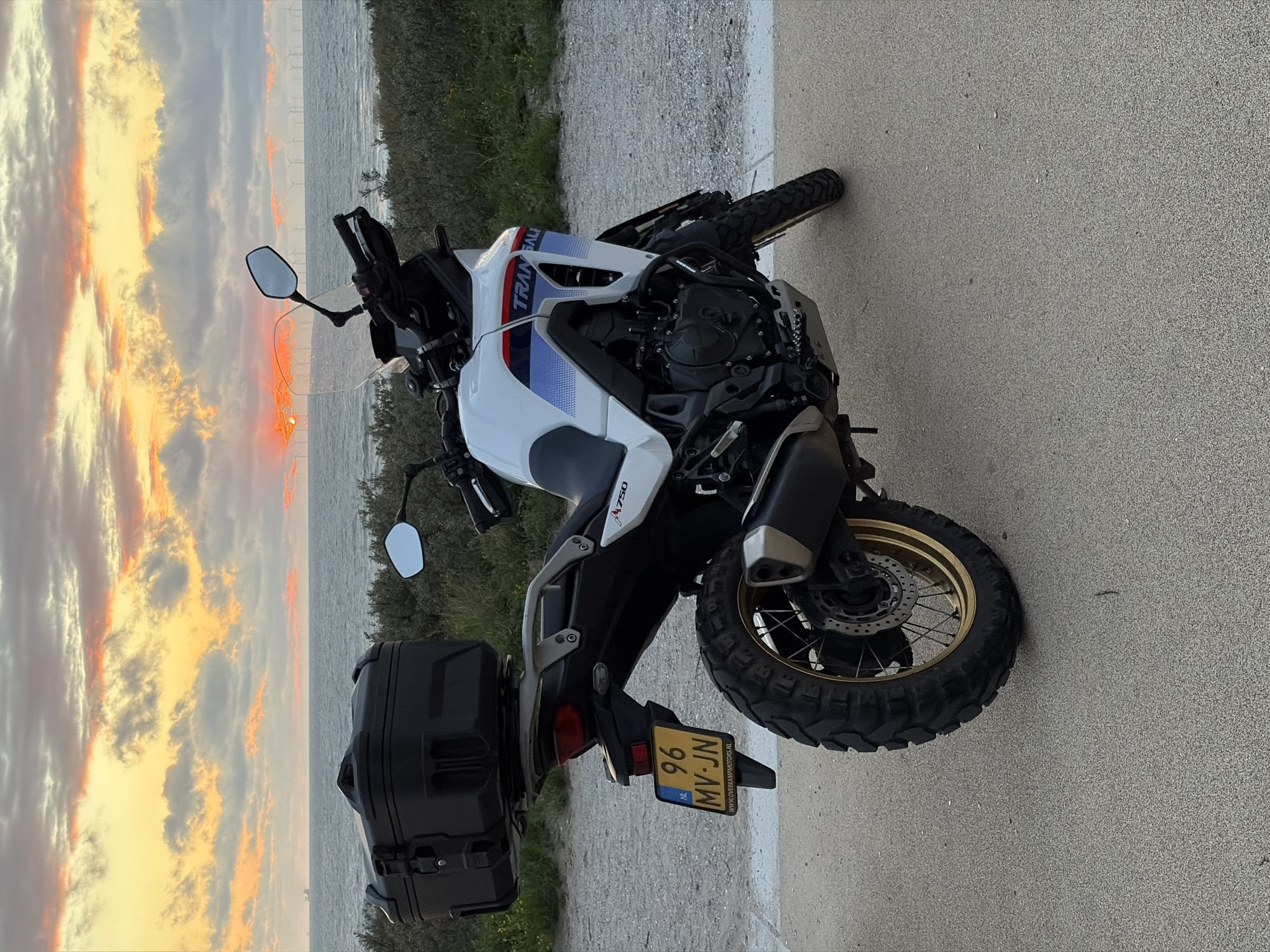 A white Honda Transalp motorcycle is parked on a paved surface next to a gravel road, with the North Sea and a wind farm visible in the background. The sun is setting over the water, casting a warm glow on the clouds. The motorcycle is equipped with a black top case and a Dutch license plate that reads "96 MV-JN". The motorcycle is facing away from the camera, with its right side prominently displayed. The environment appears to be a coastal area, possibly near Scheveningen in The Hague, Netherlands, given the presence of the wind farm and the Dutch license plate. The weather is clear, with dramatic cloud formations illuminated by the sunset.