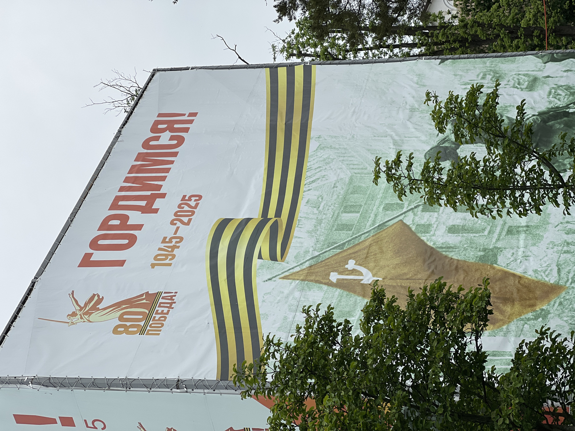A large banner is displayed outdoors, featuring the text "ГОРДИМСЯ!" (meaning "We are Proud!"), along with the dates "1945-2025" and a logo commemorating the "80th Победа!" ("80th Victory!"). The banner also incorporates a stylized image of the Saint George ribbon and what appears to be the Soviet victory banner. Part of the banner is obscured by tree branches. The location is most likely in the city of Krasnodar, Russia.