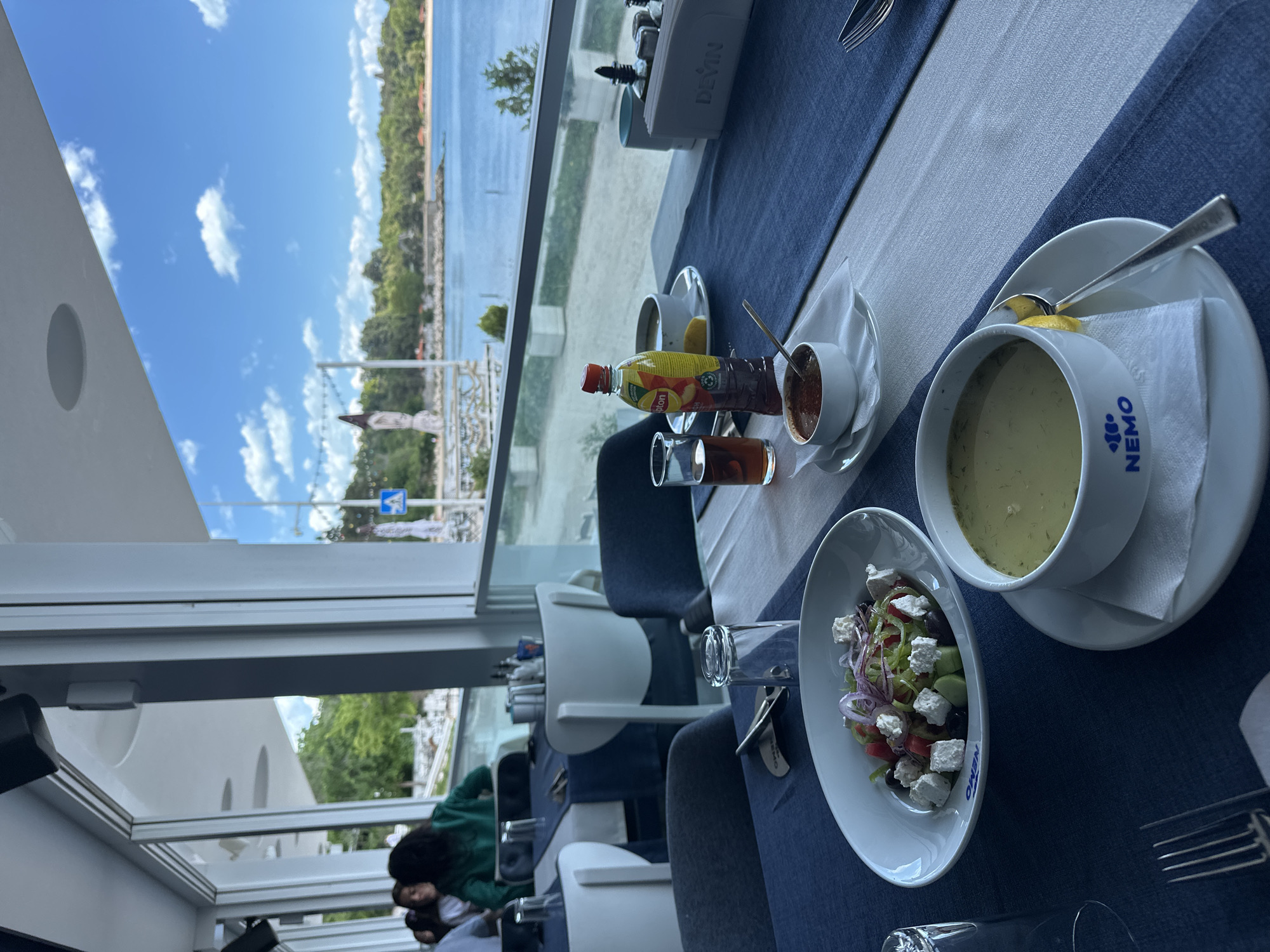 A table setting at the NEMO restaurant in Varna, Bulgaria, is arranged for dining. A bowl of soup and a salad are placed on the table alongside condiments, including a small bowl with a reddish sauce and a bottle of iced tea. The table, covered with a blue tablecloth and a white runner, faces a large window offering a view of the coast. Outside the window, the cityscape blends with the waterfront, under a sky with scattered clouds. Inside, people are seated at adjacent tables, visible in the lower-left corner of the frame.