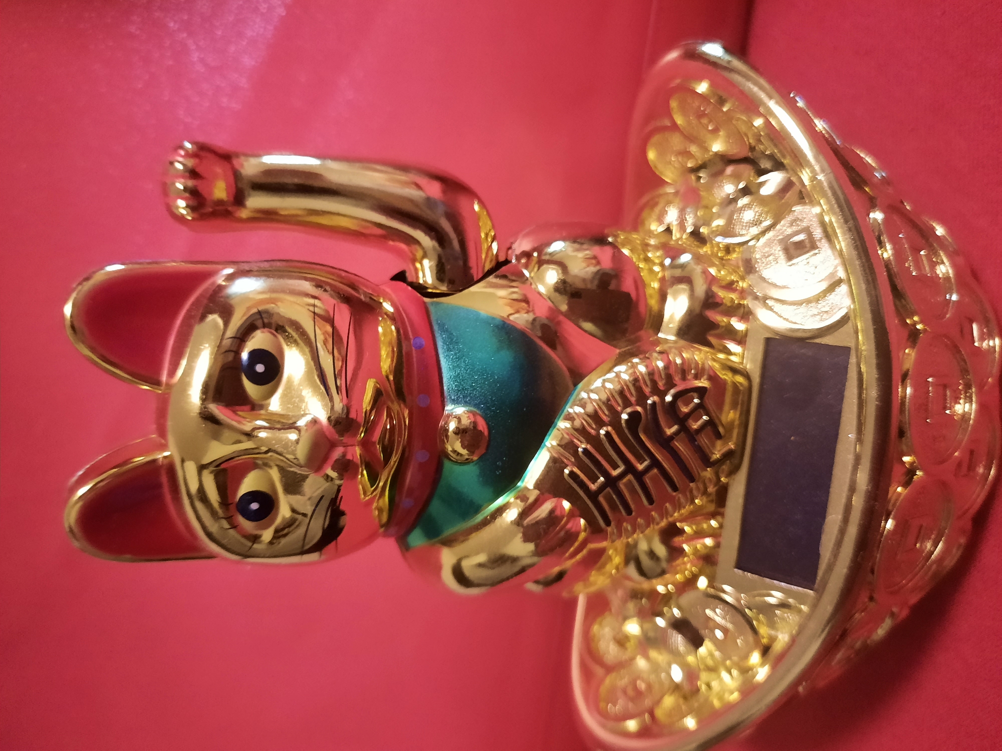 A golden, fortune cat figurine is centered in the frame against a vibrant pink backdrop. The figurine depicts a cat with one paw raised, holding what appears to be a small solar panel on a base adorned with golden coins. The cat wears a green and red collar, and a golden charm hangs from its neck. The figurine is placed on a golden dish filled with more golden coins. The lighting is bright and casts reflections on the metallic surface of the figurine. The image is sharp and detailed, with a shallow depth of field that blurs the background.