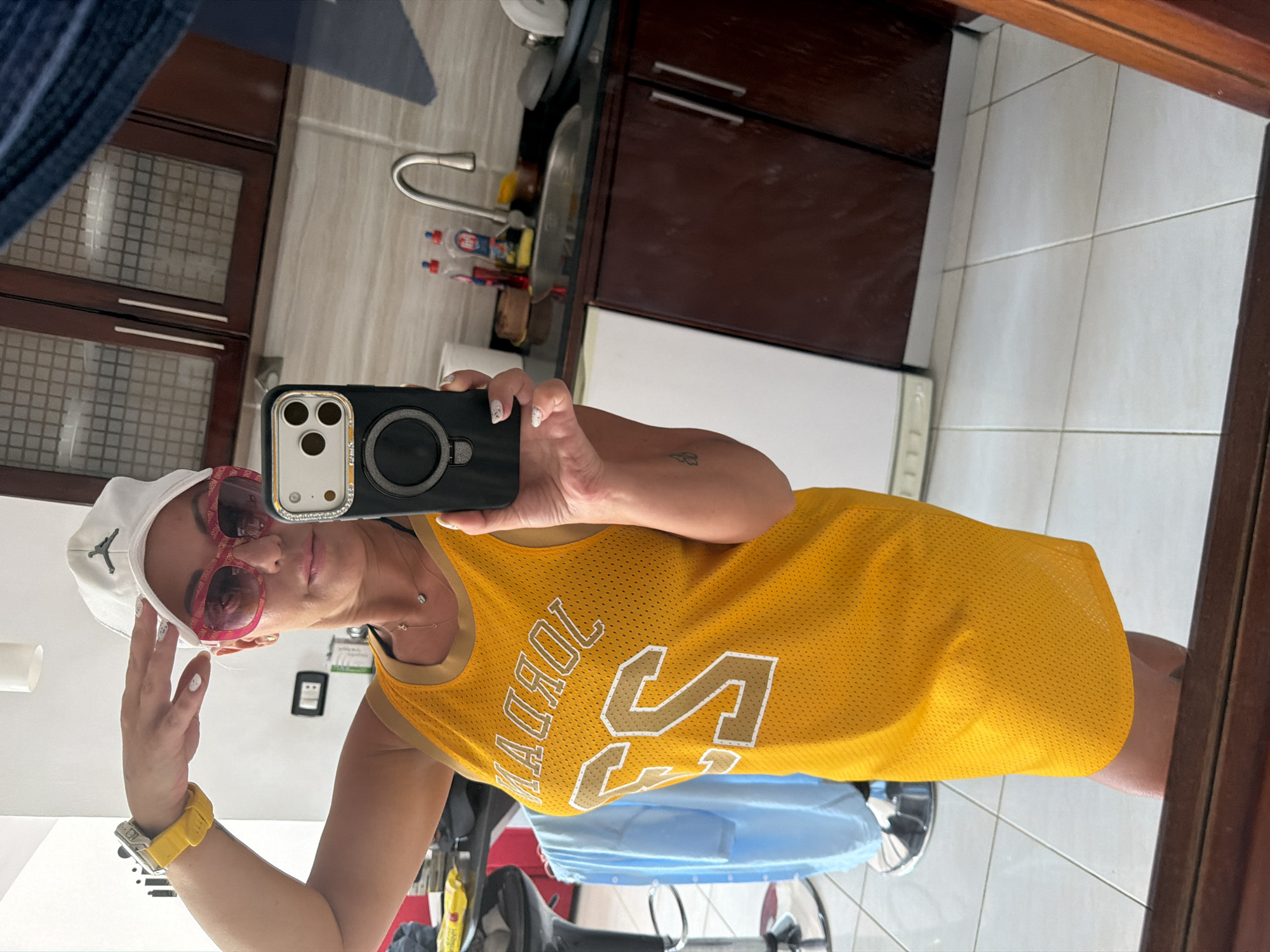 In Sharm El Sheikh, Egypt, a woman stands in a kitchen, taking a mirror selfie. She wears a white baseball cap with a black Jumpman logo, large pink sunglasses with a patterned frame, and a yellow basketball jersey dress with "JORDAN" and the number "23" in white and gold lettering. A yellow watch is visible on her left wrist. Her right hand is raised to her forehead, and she holds a black iPhone with a silver camera surround. A thin silver necklace with a small pendant is visible around her neck. The kitchen features dark brown cabinets, a stainless steel sink with a faucet, and tiled walls and floors. A blue shirt hangs on a stool in the background.