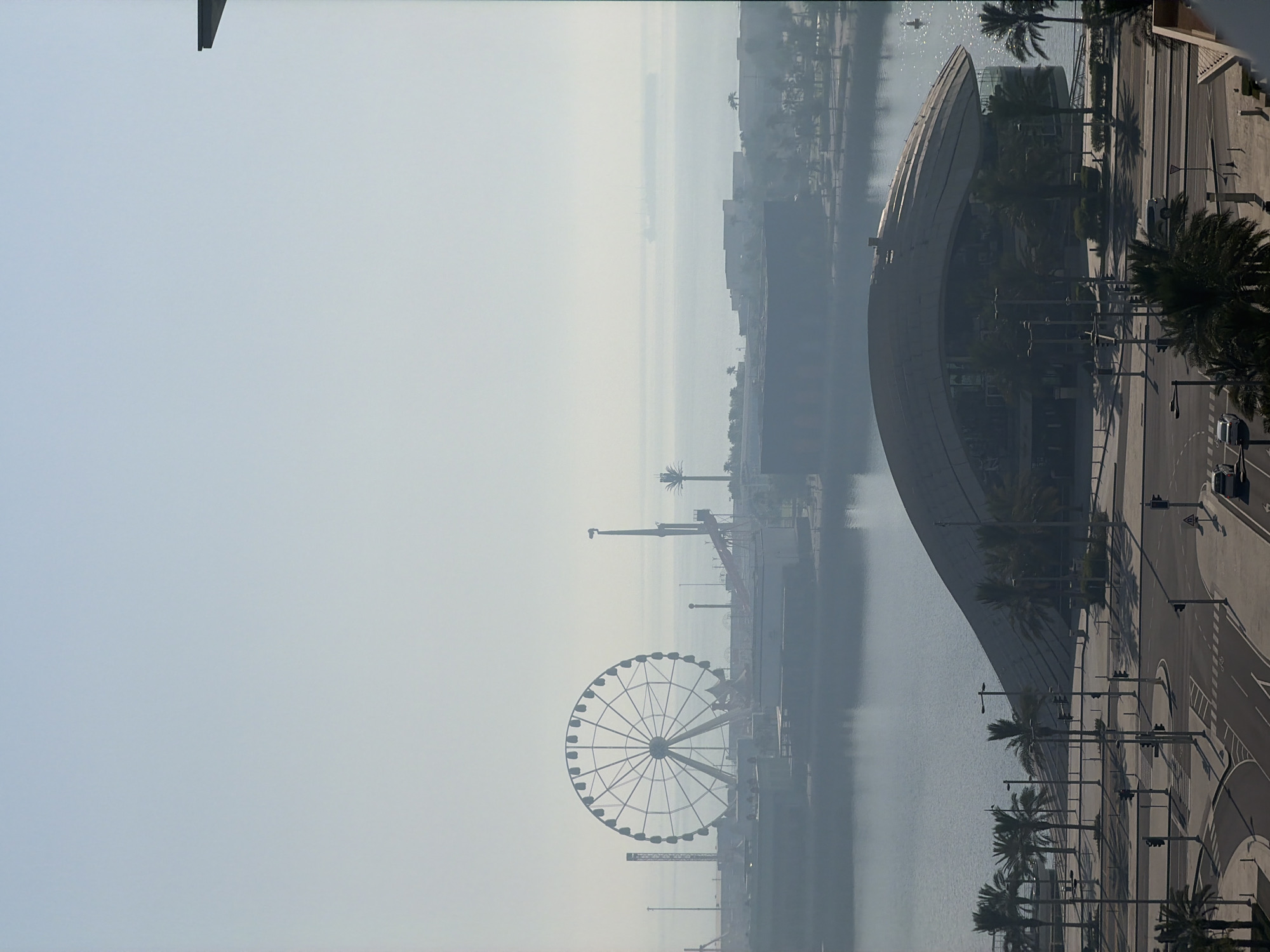 This media file captures a hazy, likely early morning or late afternoon, view of a waterfront area featuring a large Ferris wheel and a modern, curved building. The scene is dominated by a pale sky and calm water, suggesting a serene atmosphere. Palm trees are scattered throughout the foreground and along the waterfront, hinting at a warm climate. A road with traffic signals and pedestrian crossings runs parallel to the water, with a few cars visible. The Ferris wheel stands prominently in the mid-ground, its intricate structure silhouetted against the hazy sky. In the distance, several other structures and a larger body of water with what appear to be ships can be seen, further obscured by the haze. The overall impression is one of a coastal urban development designed for recreation or tourism. No people are clearly visible. No text is visible.