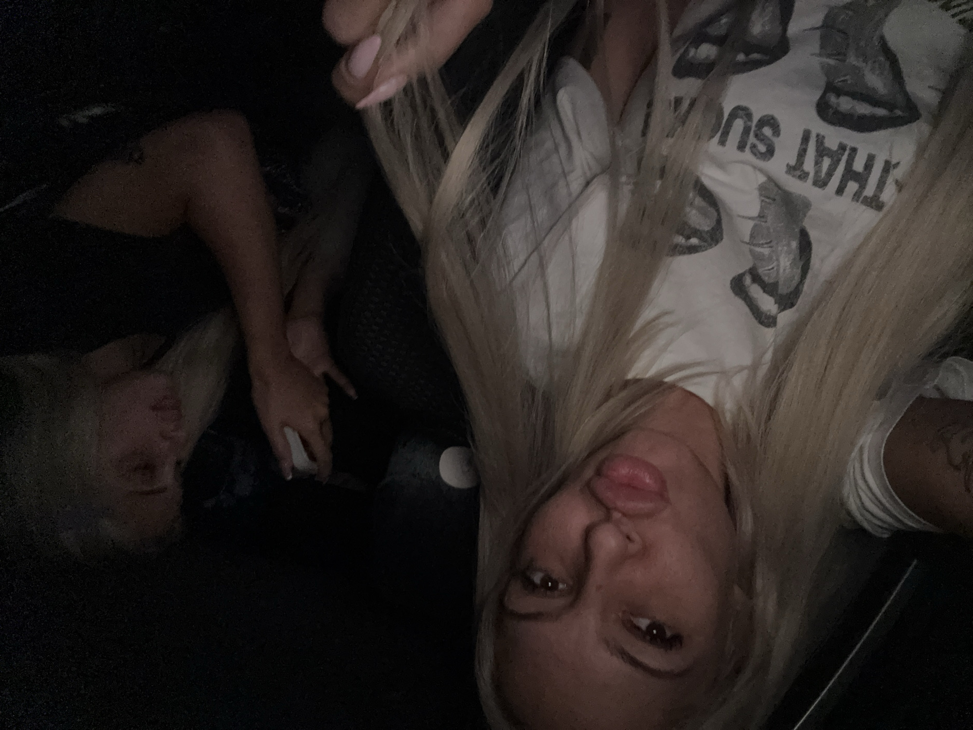 Two women are present in the image. One woman, positioned closer to the camera, has long blonde hair and is wearing a white t-shirt with a graphic print. The other woman, further away, is wearing a black top. The environment appears to be indoors. There is no indication of the location in Cherna Gora, Bulgaria.