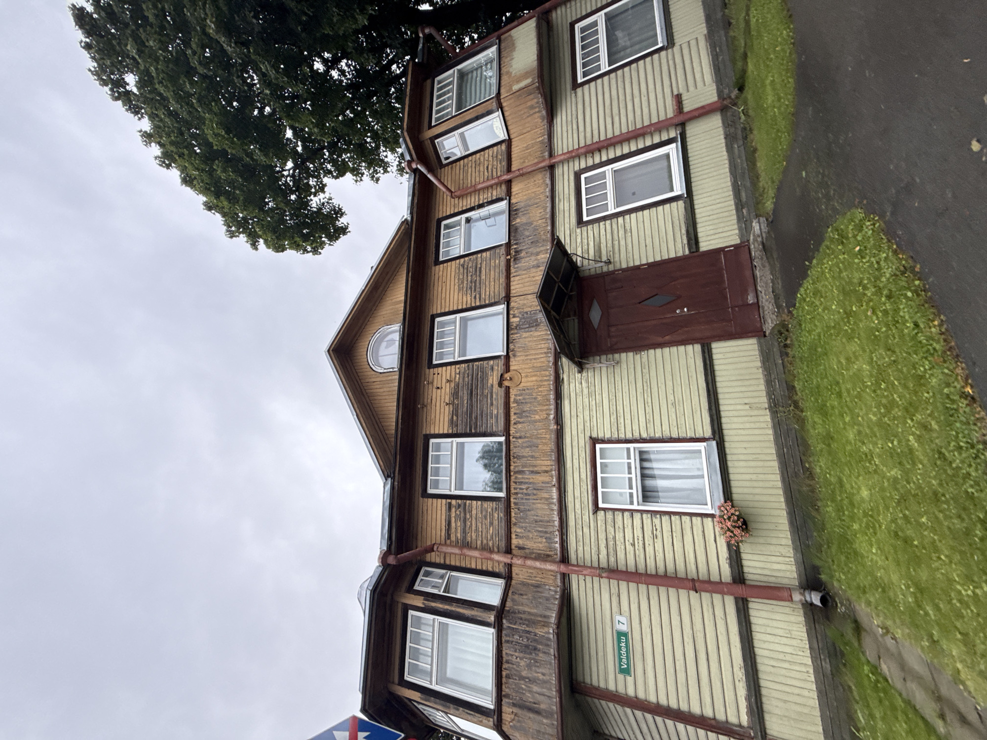 The image depicts a two-story wooden house with a pitched roof, located on Valdeku Street, house number 7, in Tallinn, Estonia. The house has a faded yellow siding on the lower level and weathered brown wood on the upper level. It features several white-framed windows, some of which are bay windows on the upper floor. A dark red wooden door is visible on the lower level, partially obscured by a small awning. Red drainpipes run down the sides of the house. Green grass surrounds the property, with a dark asphalt path leading to the door. A hanging flower basket with pink flowers is attached to the exterior wall above the lower-level windows. A street sign that reads "Valdeku 7" is mounted on the wall to the left of the door. A large tree with dark green foliage is present to the right of the house, partially obscuring the sky. The sky is overcast and grey. The image is taken from a low angle, looking up at the building. There are no people visible in the image.