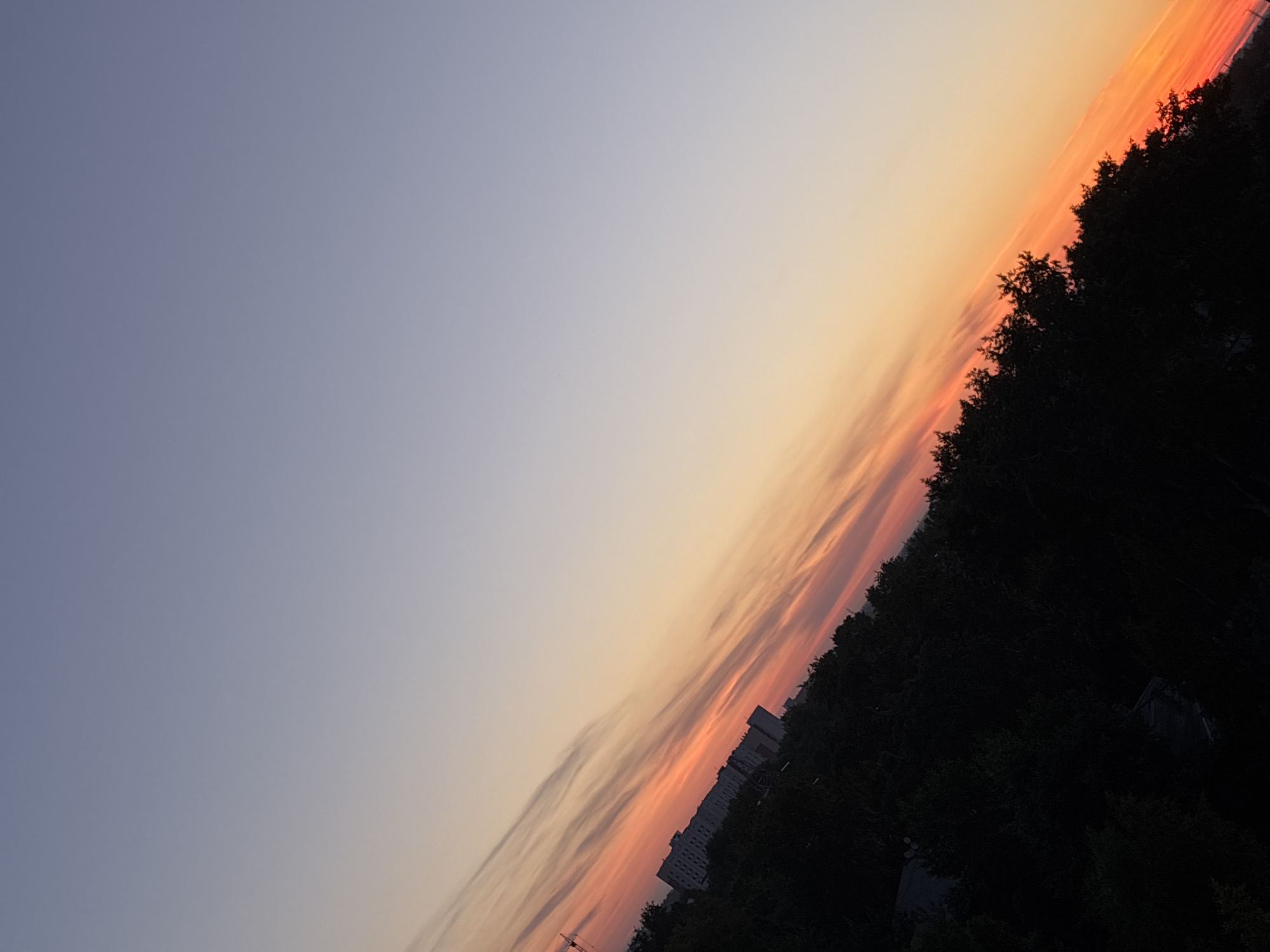 The image captures a diagonal view of a cityscape at sunset. The sky transitions from a pale blue at the top to a warm gradient of orange and pink hues near the horizon, streaked with wispy clouds. In the lower left portion of the image, a dense line of dark trees forms a silhouette against the vibrant sky. Behind the trees, a long, multi-story building is visible, suggesting an urban environment. A construction crane is also partially visible on the far left, further indicating a developed area. The overall impression is a serene urban sunset scene.