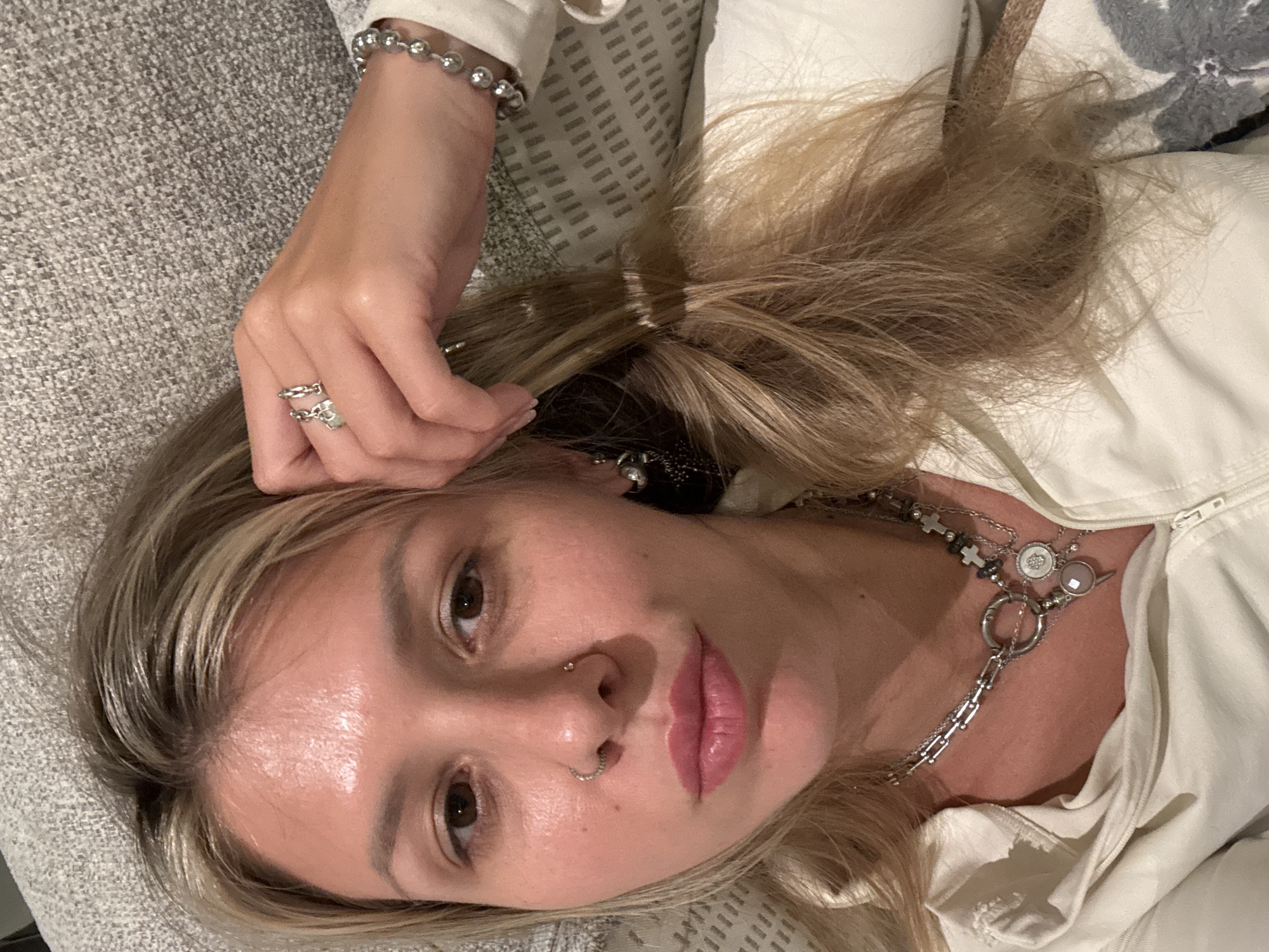 A young woman with blonde hair is lying on a couch, resting her head on her hand. She is wearing a white hoodie and several pieces of jewelry, including rings on her fingers, a bracelet on her wrist, and layered necklaces. The woman has a nose ring and earrings. She is looking at the camera, and her lips are painted pink. The background of the image shows a textured gray couch and a white pillow with a gray pattern. The lighting is soft, suggesting an indoor setting.