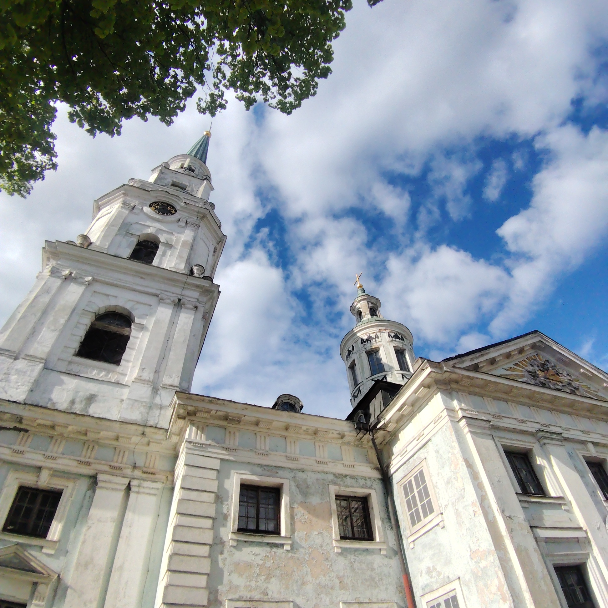 Low-angle view of the Peter's Church in Riga, Latvia, showcasing its bell tower with a clock and a green spire, as well as a portion of the main building with architectural details like windows and decorative facades. The sky is partly cloudy, with blue patches interspersed with white clouds. The upper left corner of the image features tree foliage. The building's paint appears weathered in certain areas.