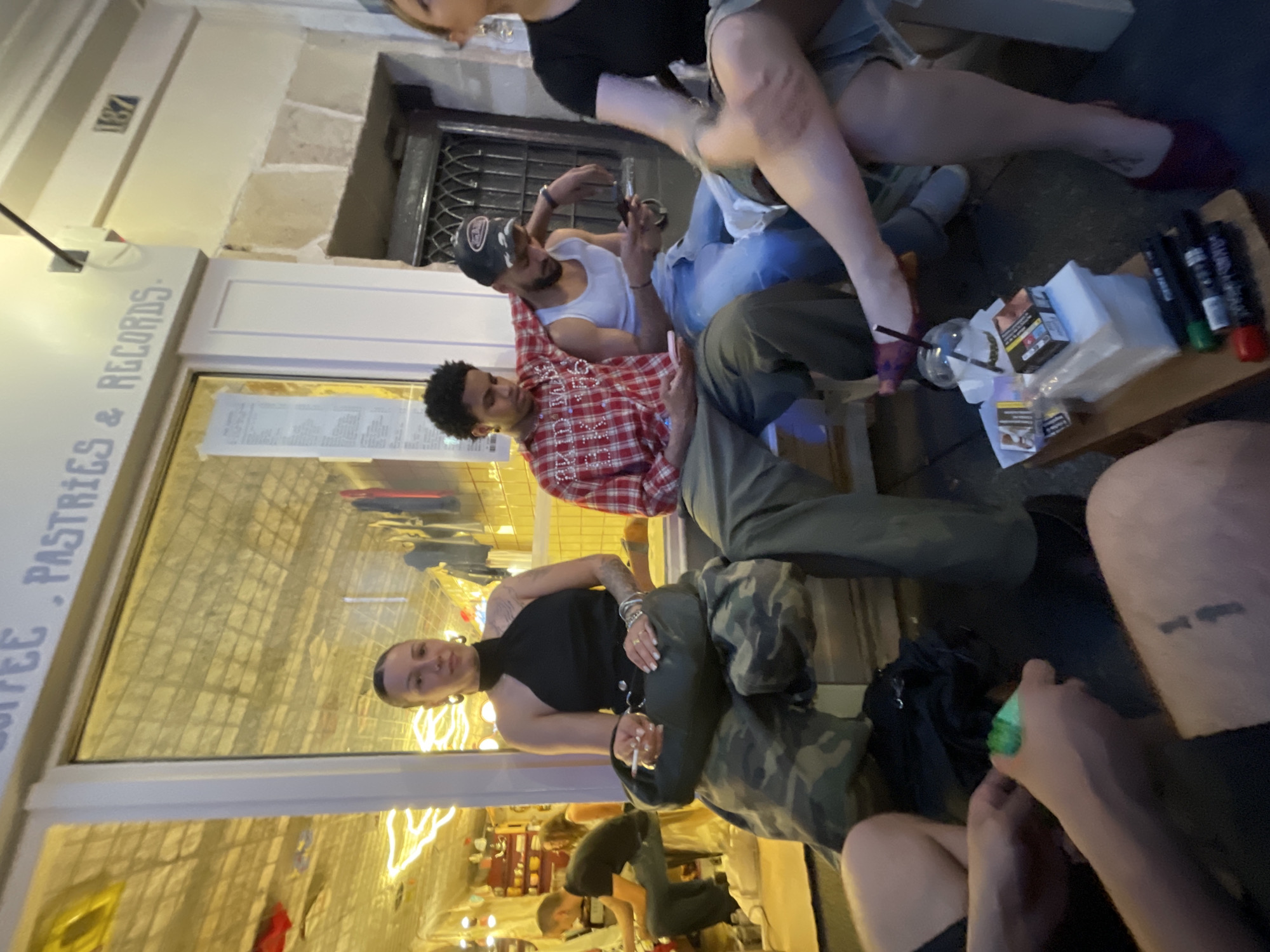 The photograph shows a group of people sitting and standing outside a business in Paris, France, with "COFFEE . PASTRIES & RECORDS" signage. A woman in a black halter top and camouflage pants sits with a cigarette in hand. Another person in a red plaid shirt looks at their phone. A woman sits on the table with her legs dangling, while another person wears a tank top and a hat. On the table are cigarettes, markers, and papers. The storefront window reflects an interior scene of a cafe or bar. The number "187" is visible on the wall.