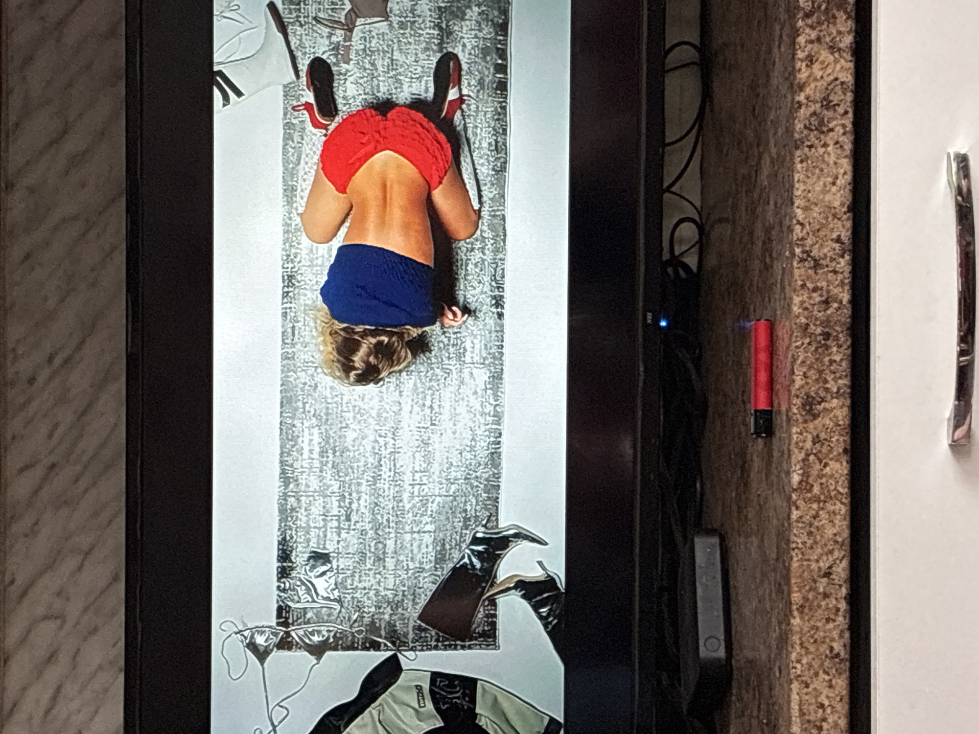 A photograph shows a television screen displaying an image of a woman lying face down on a patterned rug. She is wearing red shoes, a red top, and blue shorts. Beside her are various clothing items, including a white and black jacket, bikini tops, and black boots. The television is positioned next to a cabinet with a granite-like pattern. A red object is adhered to the side of the cabinet. The photograph was presumably taken in an interior space, but the precise location in Varna, Bulgaria cannot be determined from the visual information provided.