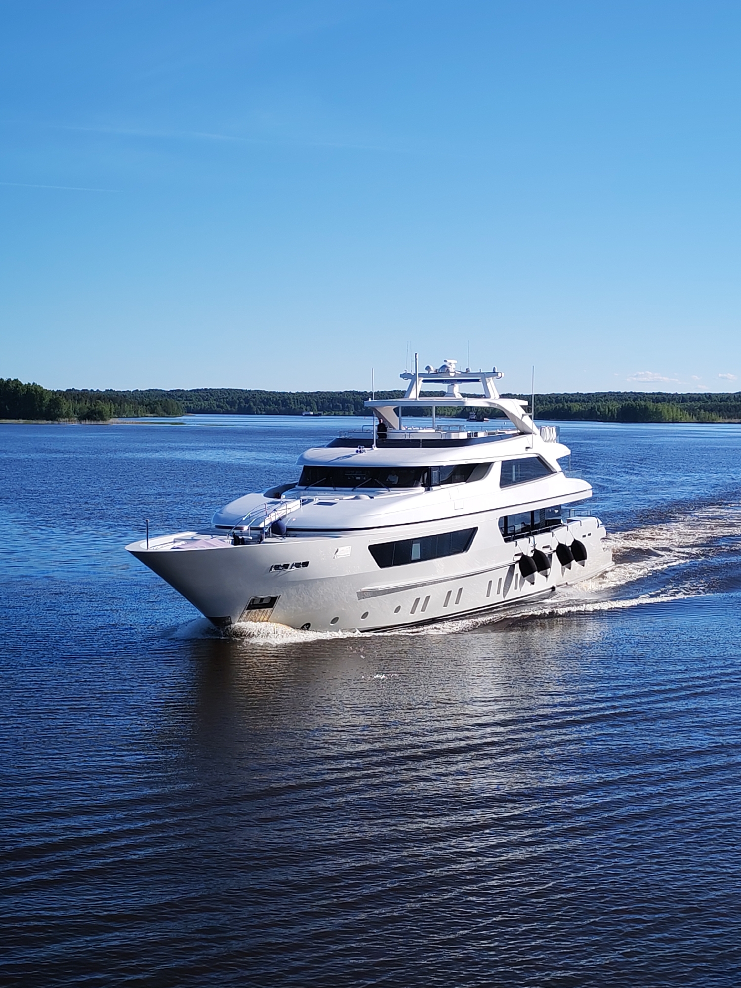 A large, modern multi-deck motor yacht, predominantly light grey and white, navigates calm, deep blue water in Novodevich'e, Russia. The vessel is positioned centrally, moving from right to left, generating a distinct white bow wave at its front and a frothy wake behind its stern. Several black cylindrical fenders are visible along the starboard side of the yacht. The yacht features multiple large rectangular windows on its main and upper decks, and an array of communication and navigation equipment is mounted on the uppermost deck. In the distant background, a dense, verdant forest lines the shores on both sides of the wide waterway. The sky above is clear and bright blue, with a few faint white contrails present. The scene is brightly illuminated by direct sunlight. No individuals are clearly identifiable aboard the yacht or in the surrounding environment.
