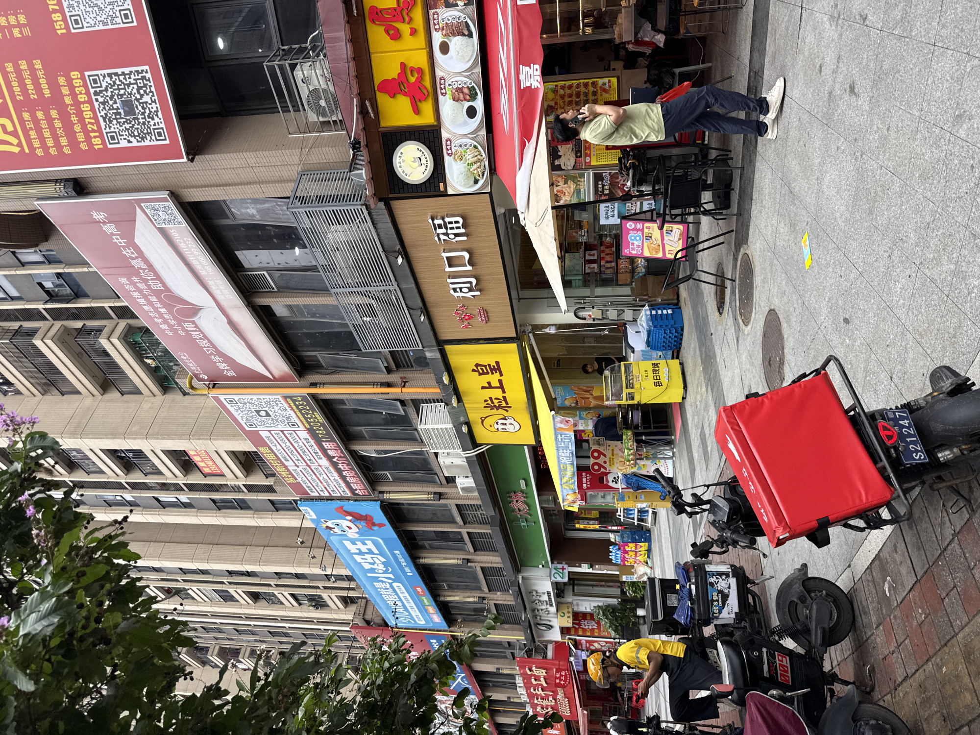 In a bustling Haizhu District street scene in China, a delivery person in a yellow shirt and black apron sits on an electric scooter, looking at a phone. Another electric scooter with a large red insulated delivery box is parked nearby. A man in a green shirt and dark pants stands on the sidewalk, talking on his phone. The street is lined with businesses, including a restaurant with a red awning and a food stall displaying prices and food items. Buildings in the background are multi-story with balconies. The sidewalk is paved with gray tiles, and several manhole covers are visible. A tree with green leaves and some purple flowers overhangs the scene from the left.