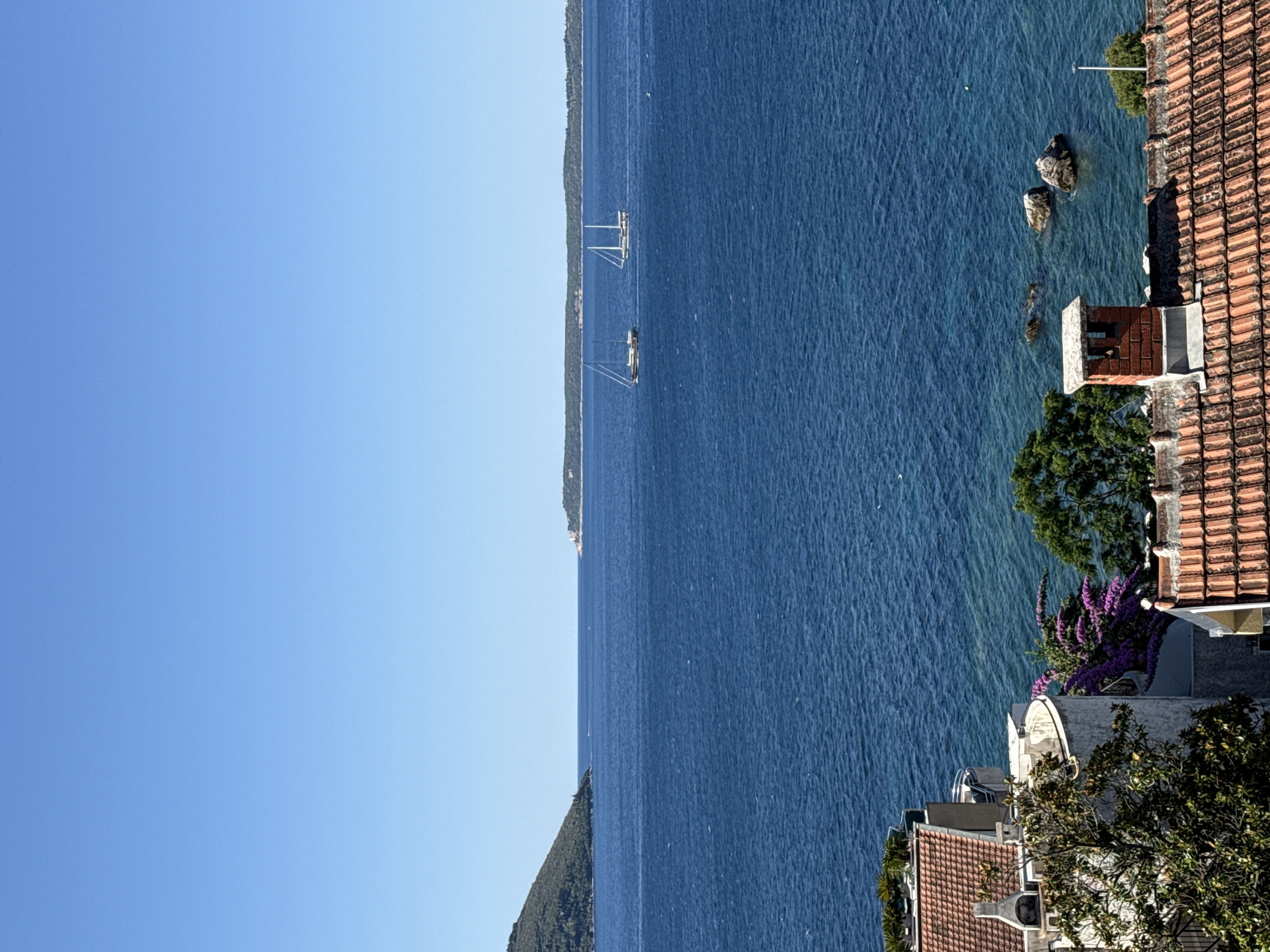 The image shows a view of the Bay of Kotor near Herceg Novi, Montenegro. In the foreground, there are terracotta tiled roofs of houses, one with a brick chimney. Further down, a white building with a curved balcony is visible, adorned with vibrant purple bougainvillea and lush green trees. The middle ground is dominated by the deep blue, choppy waters of the bay, with several rocks breaking the surface near the shore. Two sailboats are visible in the distance on the water, with one appearing slightly ahead of the other. The background features a green, tree-covered hillside on the left, a distant coastline with structures on the right, and a clear, bright blue sky above. The sun is high, indicating it is likely daytime.
