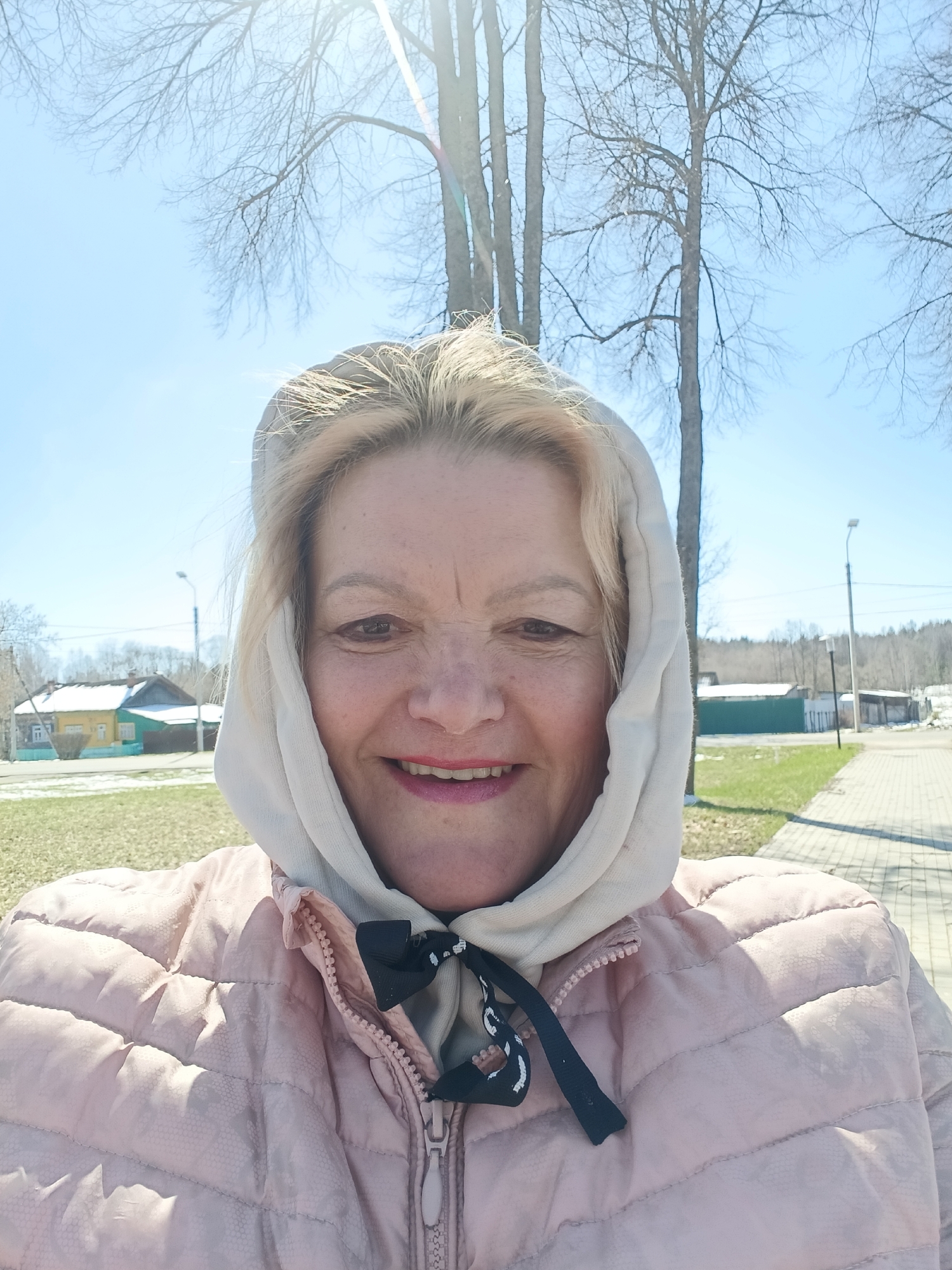 A cheerful, blonde-haired woman is captured in a sunlit outdoor selfie, smiling broadly at the camera. She appears to be middle-aged and is wearing a light-colored hooded top, possibly a hoodie, tied under her chin with a black and white patterned ribbon, and a light pink quilted jacket. Her face reflects a happy and relaxed mood.
The background reveals a clear, bright blue sky with significant lens flare from the sun in the upper center, suggesting a sunny day, likely around midday or early afternoon. Tall, bare trees, characteristic of late winter or early spring, stand against the sky. Patches of green grass are visible on the ground, interspersed with areas of brown earth and remnants of snow, particularly on the roofs of houses in the distance to the left. A paved pathway runs along the right side of the frame, leading further into what appears to be a rural or semi-urban settlement. Streetlights are also visible in the background. The overall scene indicates early spring weather, cool but sunny. The location context indicates Красный Маяк, Russia. No visible text is present in the image.