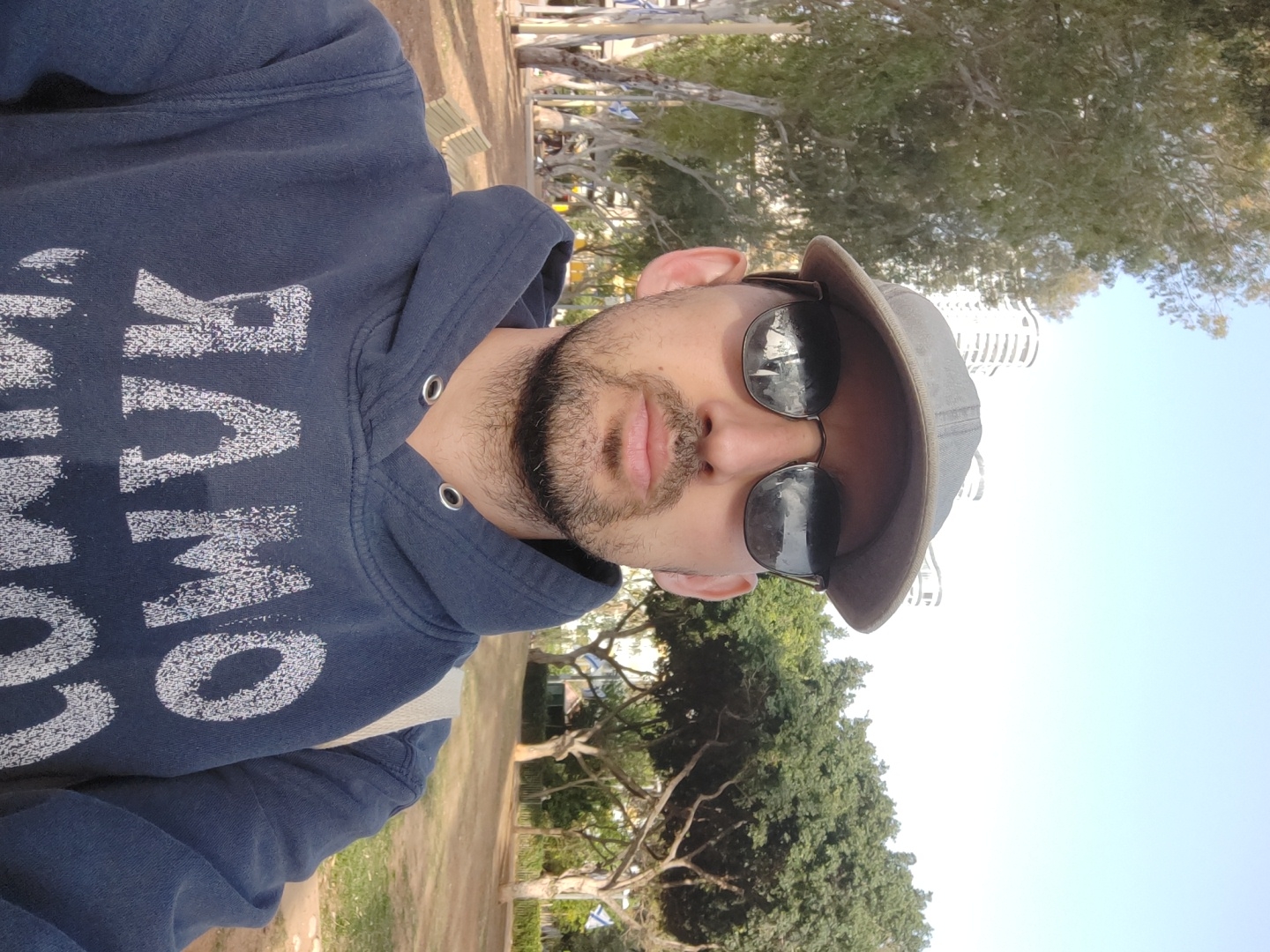 A man with a beard and sunglasses wearing a cap and a blue hoodie with white lettering is standing in a park. Behind him, trees, grass, benches, and a light blue sky can be seen. In the background, buildings can be spotted, which suggests the man may be in a city park. Given the user's request for the location to be specified as Tel-Aviv, Israel, the scene is likely a park in that city.