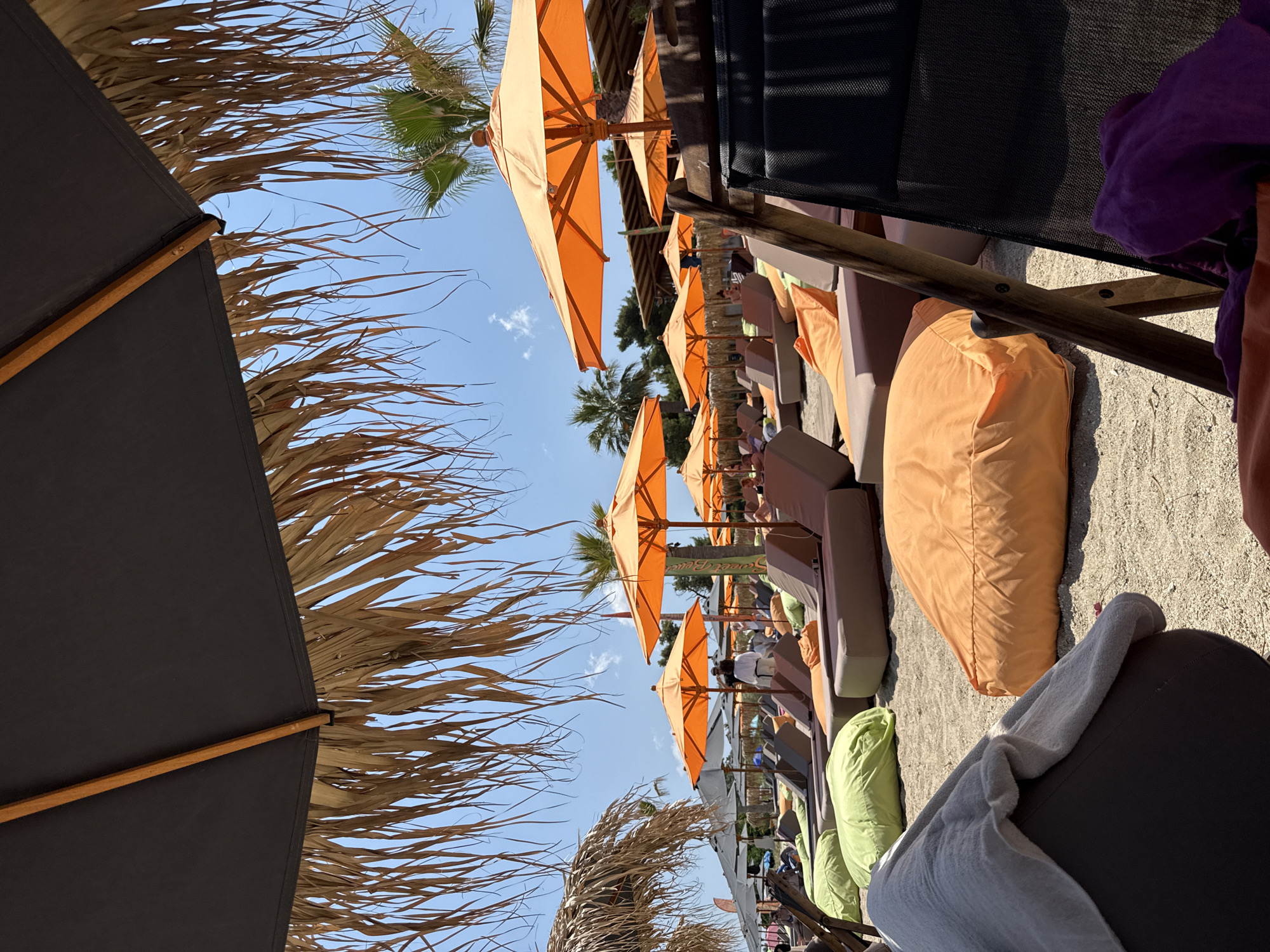 The image depicts a sun-drenched beach in Pallini Municipal Unit, Greece, featuring rows of lounge chairs and beanbag-style seating arranged on sandy ground. Several orange umbrellas provide shade, some with thatched edges. Palm trees and other foliage are visible in the background, under a clear blue sky with sparse clouds. The scene suggests a resort or beach club environment, with lounge areas set up for relaxation. The perspective is from a seated position, looking out over the beach setup.