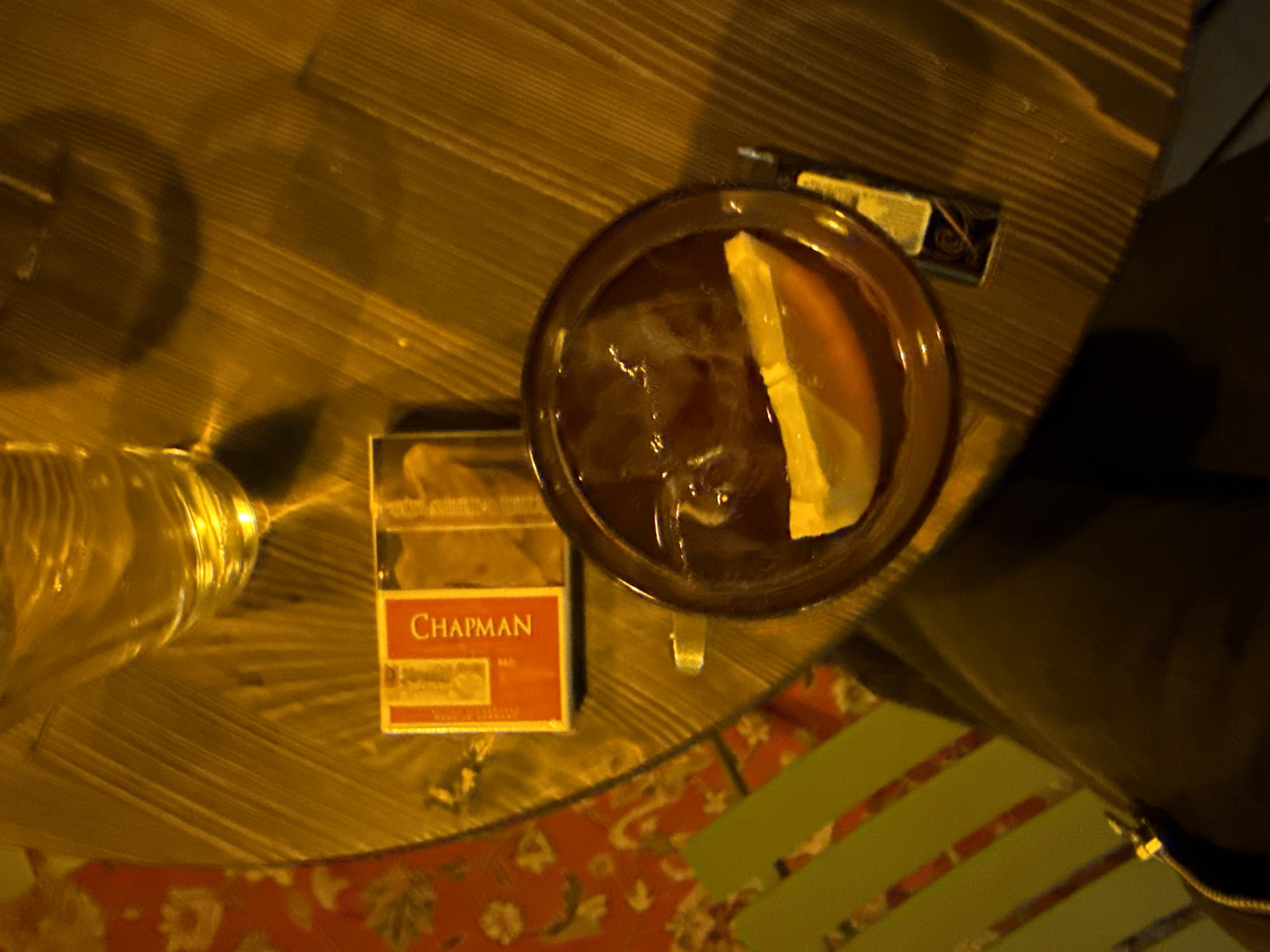 A high-angle shot depicts an overhead view of a dimly lit table setting, possibly in Yerevan, Armenia. A pack of Chapman cigarettes is positioned beside a glass containing a dark liquid with ice and citrus, possibly a cocktail. A matchbox is seen near the cocktail glass. A glass of water is also visible on the table. The table appears to be made of wooden planks with a dark varnish. A portion of a chair with a red floral pattern and green stripes is visible at the bottom.