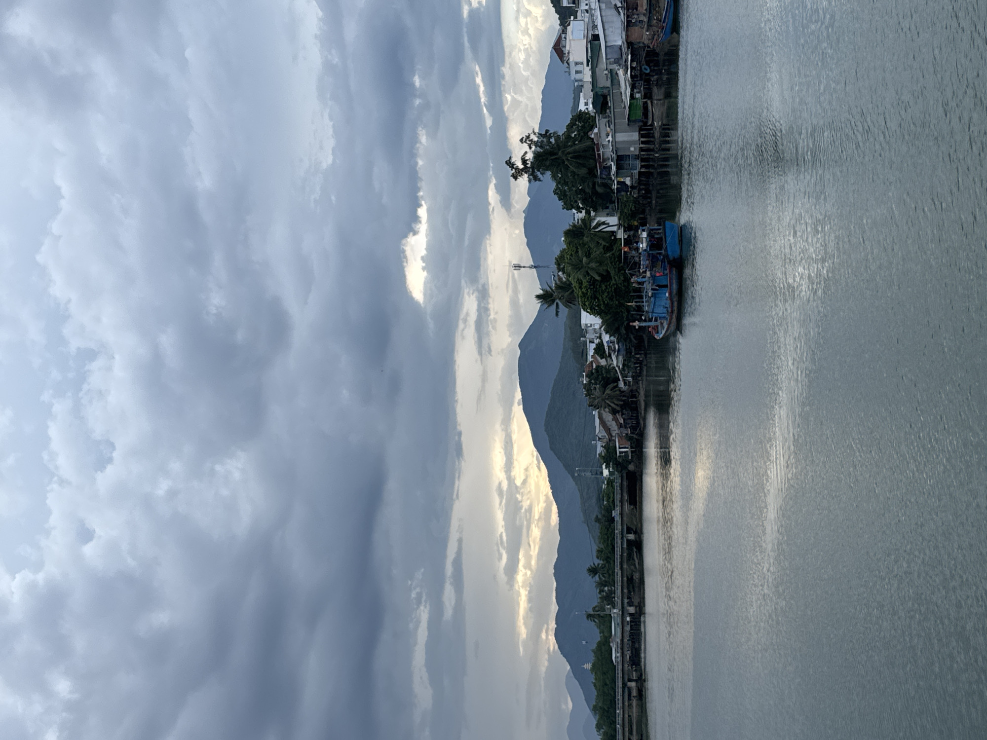 This image depicts a tranquil river scene in Nha Trang, Vietnam, likely near the Cồn Ngô district, characterized by a slightly overcast sky and calm waters. In the foreground, the broad, gray river reflects the muted light. Along the far bank, a settlement of stilt houses and smaller buildings lines the water's edge, with lush green trees interspersed among them.
Dominating the middle ground is a long, concrete bridge, possibly the Cồn Ngô bridge, with vehicles visible crossing it. Several blue fishing boats are moored along the river, one prominently featuring a tall antenna. Beyond the settlement, a range of green, rolling mountains forms the backdrop, their peaks softened by the distance and atmospheric conditions. The sky above is filled with dramatic, heavy gray clouds, with patches of lighter sky and hints of sunset peeking through the gaps, suggesting either dawn or dusk. The overall atmosphere is serene and picturesque, typical of a coastal Vietnamese town.