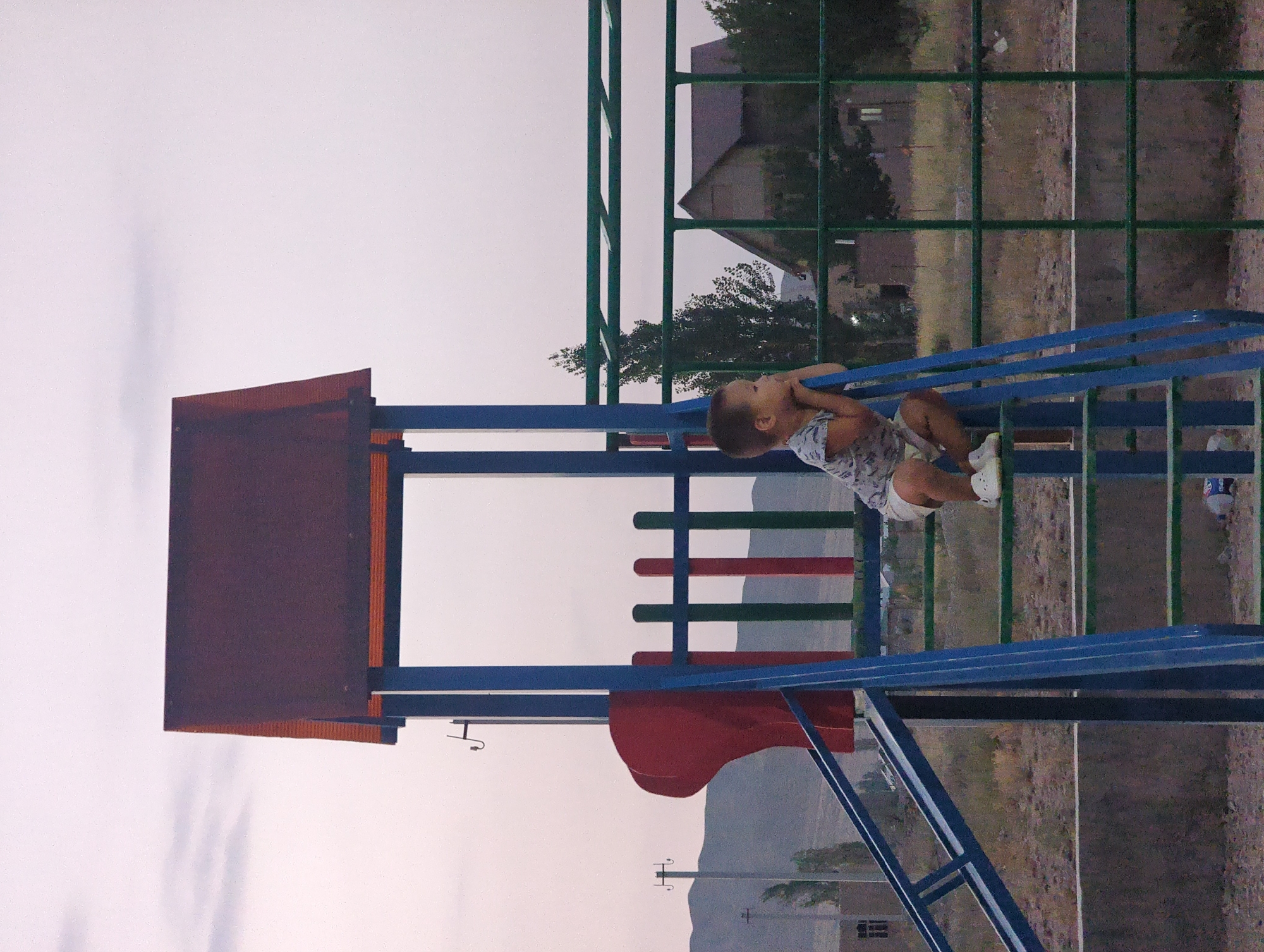 In the village of Uch-Dobo, Kyrgyzstan, a young boy sits on the stairs of a blue and green playground structure, his chin resting on his hands as he looks off to the side. He wears a light-colored t-shirt with a pattern and white shorts, along with white slip-on shoes. The playground features a red slide and a red structure above the stairs. In the background, there are trees, a building with a dark roof, and mountains in the distance under a cloudy sky. The ground around the playground is dirt and scattered dry grass.