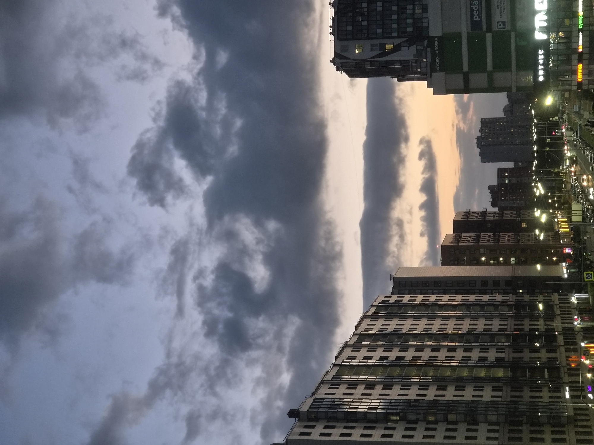 The image captures a twilight scene in an urban environment, likely a city in Russia given the Cyrillic text on signage. The sky is dominated by dramatic, dark clouds with hints of orange and pink from the setting sun peeking through. In the foreground and midground, several tall, modern apartment buildings are visible, their windows lit from within. Some buildings feature extensive glass facades that reflect the sky. A wide street with moderate traffic runs horizontally across the lower portion of the image, illuminated by streetlights. Several commercial establishments are located at the ground level of some buildings, with glowing neon signs advertising their services, one of which displays the text "ВКУСНО — И ТОЧКА" and another "pedal". There are no discernible individuals in the image, and the scene appears to be devoid of specific activity other than the general flow of traffic. The overall impression is one of a bustling yet somewhat overcast urban evening.