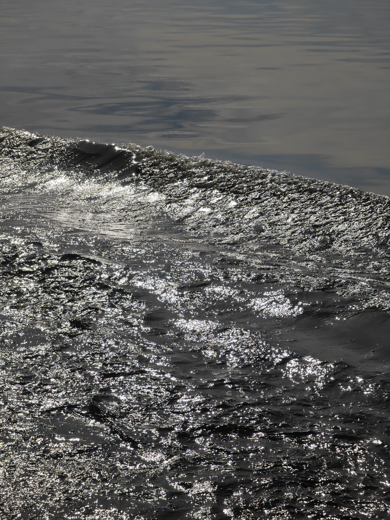 The image shows a close-up, low-angle view of dark, choppy water with the sun glinting off the surface. The foreground is dominated by the texture of the waves, which are creating a sparkling effect as the light reflects off them. In the background, the water is calmer and darker, with faint reflections of clouds or the sky. The image appears to have been taken outdoors, likely on a body of water such as a lake, river, or sea. There are no people, man-made objects, or specific landmarks visible that would pinpoint the location to Krivlyak, Russia.