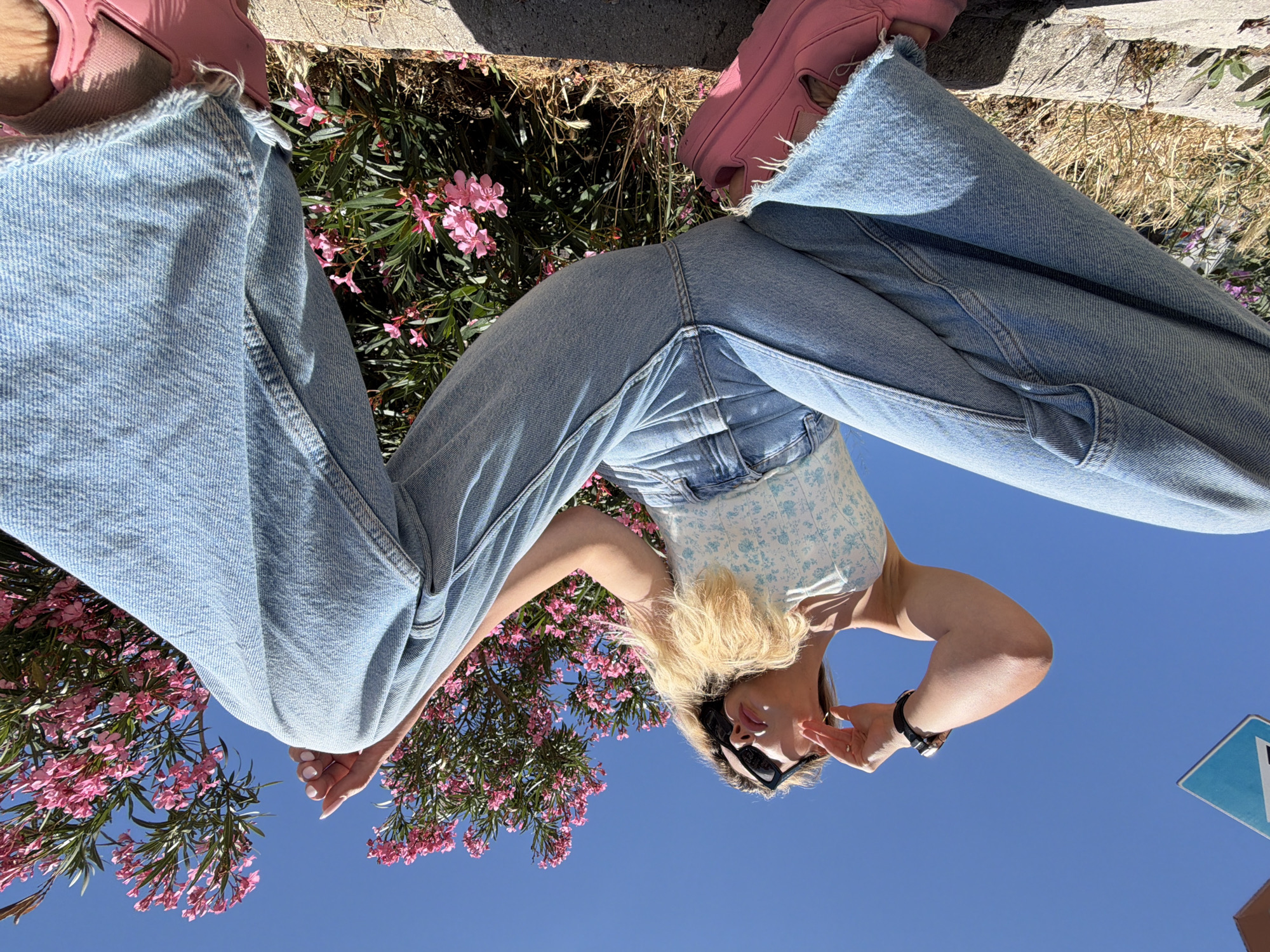A low-angle outdoor photograph depicts a woman posing in the city of Budva, Montenegro, under a clear blue sky. The woman has blonde hair and wears black sunglasses, a white strapless top with a light blue floral pattern, light blue wide-leg jeans with raw-cut hems, and bright pink platform-style shoes. A dark watch is visible on her left wrist, and her fingernails are painted white. She is positioned with her legs splayed wide towards the viewer, her left hand near her face, appearing to adjust her sunglasses, and her gaze directed towards the left side of the frame. The background features lush green bushes densely covered with vibrant pink flowers, consistent with Oleander, particularly prominent on the right. A partial blue and white rectangular sign is visible in the upper left corner. The foreground consists of a concrete curb or low wall alongside dry grass and dirt, indicating a sunny, possibly roadside or park-like setting.