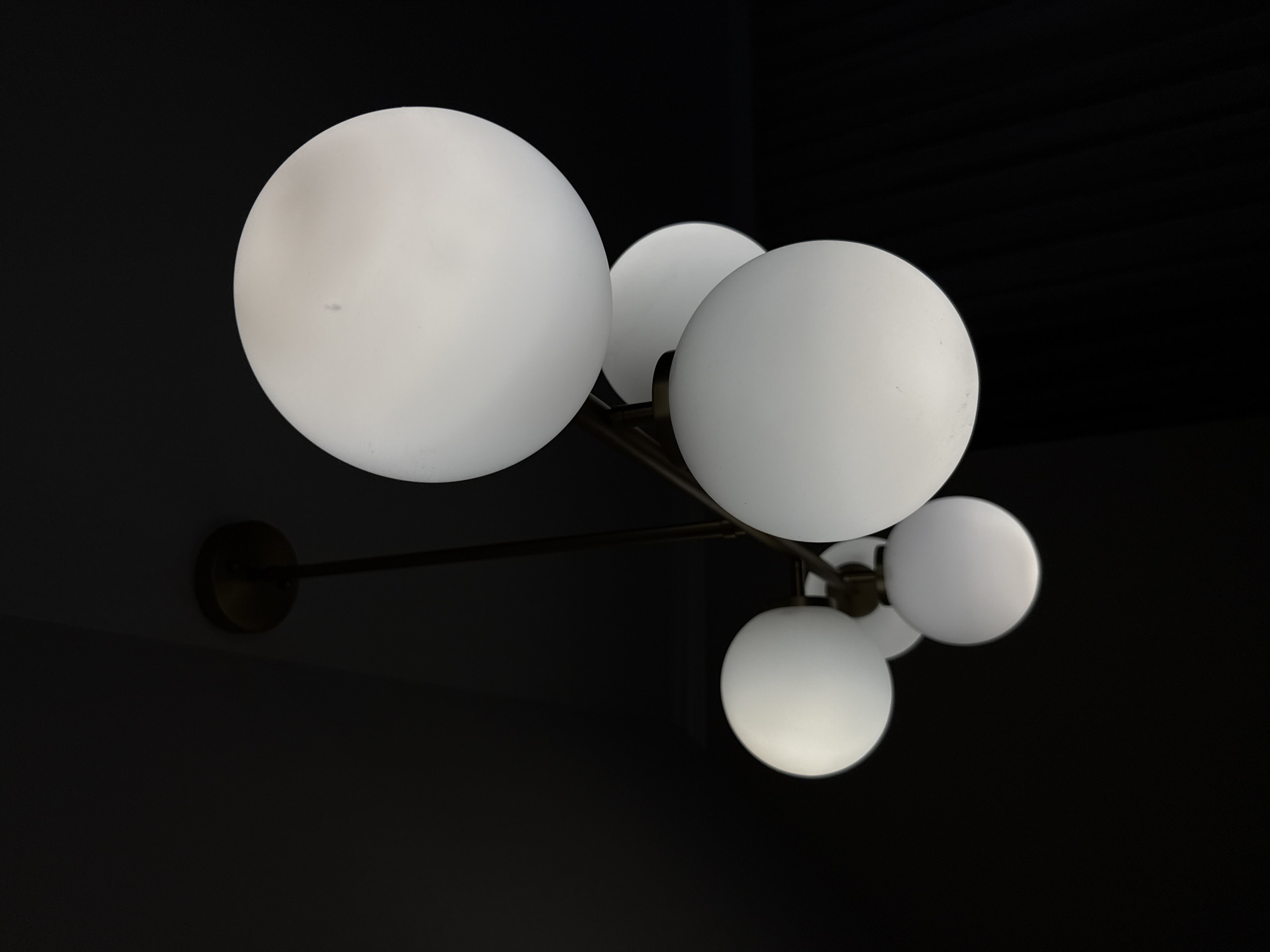 The image shows a modern chandelier with multiple frosted glass globe lights hanging from a dark ceiling. The chandelier has a minimalist design with thin metal arms. The lights are illuminated, casting a soft glow. The background is dark and out of focus, emphasizing the chandelier as the main subject. The image does not contain any recognizable landmarks or features that would indicate a specific location within Plovdiv, Bulgaria.