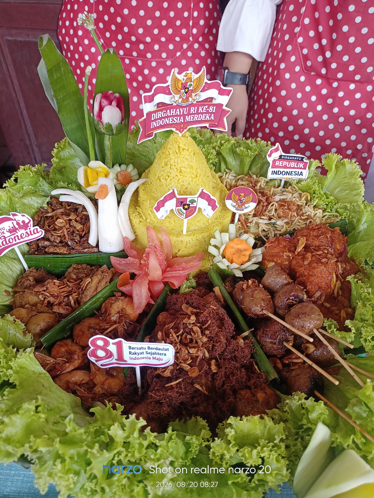 The image shows a festive Indonesian dish, likely a tumpeng, arranged on a platter. The tumpeng is cone-shaped and made of yellow rice, adorned with various food items and decorative elements. Prominently featured are Indonesian flags and emblems, suggesting a celebration of Indonesian Independence Day or a similar national event. There are also carved vegetables, fried foods, meat skewers, and fresh lettuce. Two individuals in red and white polka-dot aprons are partially visible in the background, indicating their involvement in preparing or presenting the dish. The setting appears to be indoors or a sheltered outdoor area, given the backdrop and lighting. The bottom of the image shows a watermark indicating it was taken with a "narzo realme narzo 20" on "2026/08/20 08:27", which is an anachronistic date. The overall presentation is vibrant and celebratory.