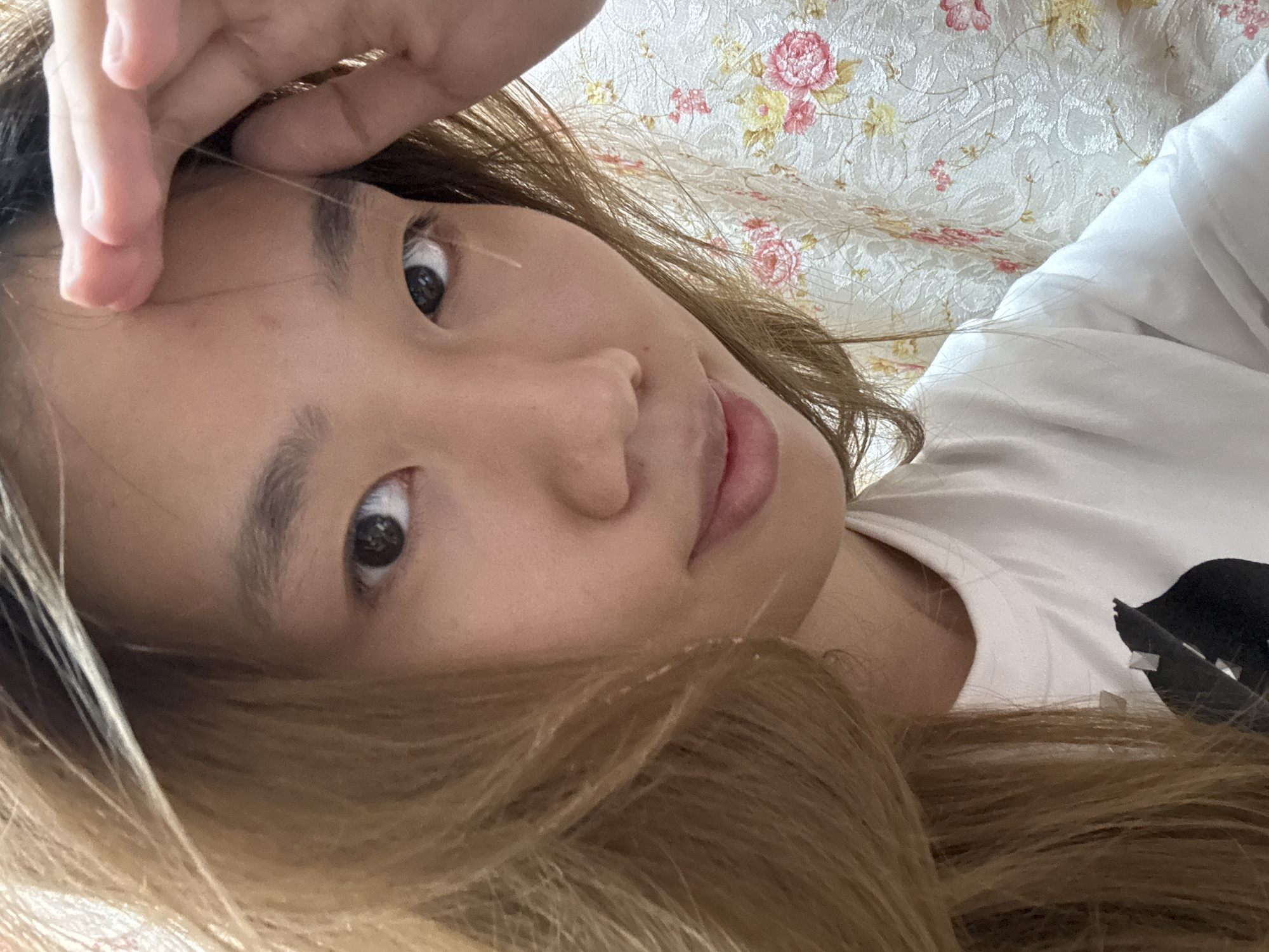 A close-up, waist-up shot features an East Asian individual with long, light brown hair that is casually styled, falling across their face and shoulders. The person is wearing a white t-shirt with a black graphic design on the left side of the chest. They are looking directly at the camera, with their right hand raised to their forehead, fingers gently touching their hairline. The background consists of a patterned fabric, likely a curtain or upholstery, with a floral design featuring pink and yellow flowers on a white textured base. The lighting is soft and diffused, creating a slightly warm tone. The setting appears to be indoors, possibly a residential space, and there is no indication of a specific location in Shapingba District, China, beyond the image content itself.