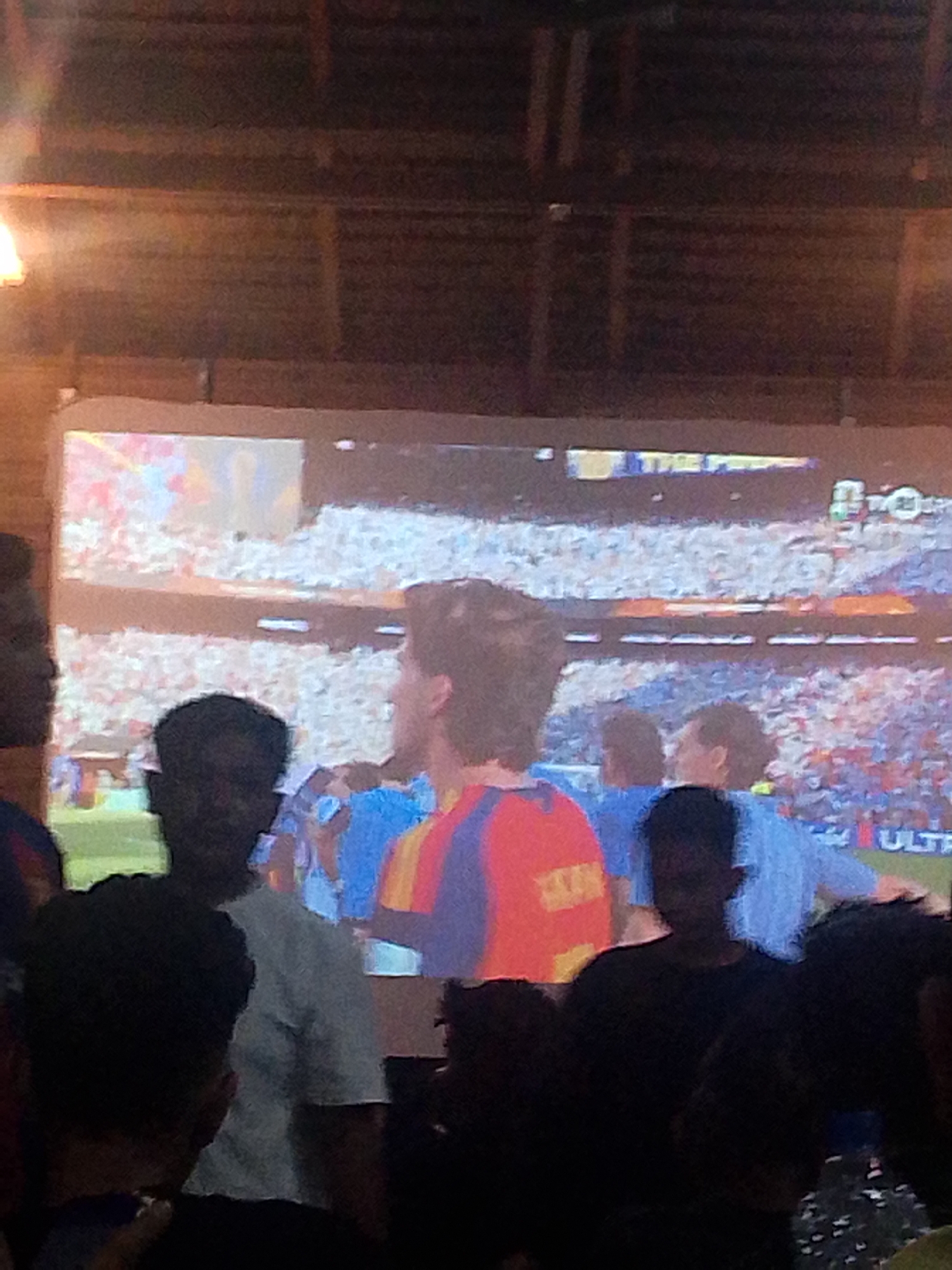 In Meunasahmee, Indonesia, a group of individuals are gathered indoors, observing a projection of a soccer match on a large screen. The projected image displays a stadium filled with spectators, with a soccer game in progress. Several people are visible in the foreground, silhouetted against the bright screen, suggesting they are seated or standing in a dimly lit area. Their faces are largely obscured, but their attention is clearly directed towards the projected game. The environment appears to be a casual gathering place, possibly a restaurant or a home, with a wooden ceiling visible in the upper portion of the image. The overall atmosphere suggests a shared experience of watching a sporting event.