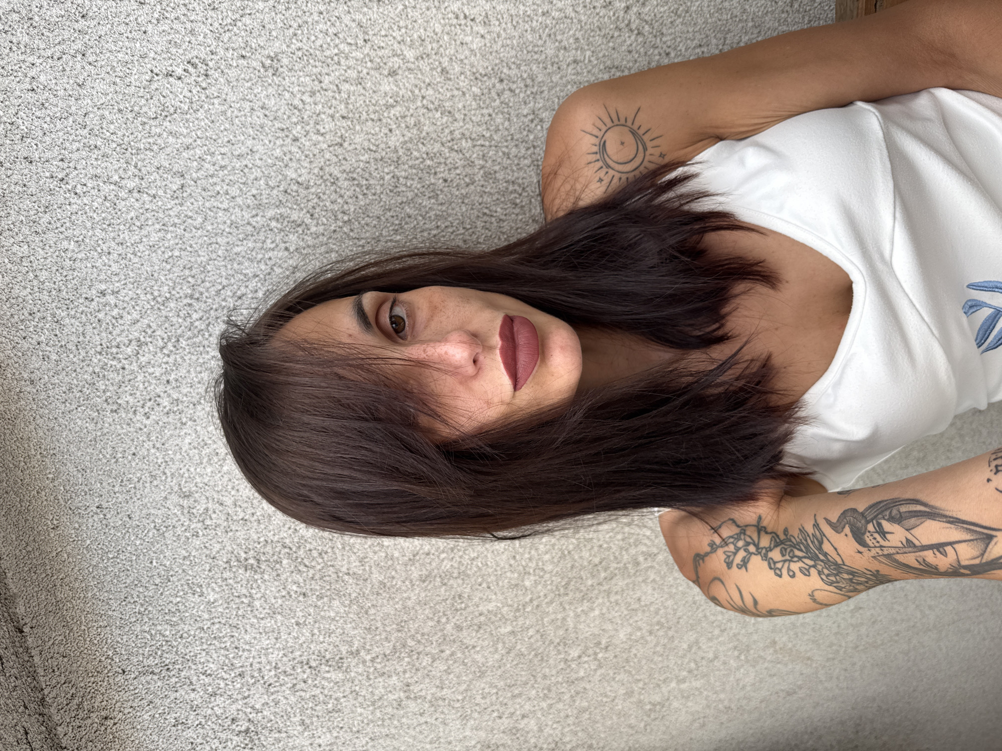 A portrait of a woman with dark hair and makeup is shown against a textured gray wall. The woman is wearing a white tank top. She has tattoos on her left arm and shoulder. The image is taken from a medium shot, with the woman's face in the center of the frame. The background is out of focus, drawing attention to the woman. The lighting is natural, and the overall mood is casual and relaxed. The image does not provide any specific information about the location in the city of Pleven, Bulgaria.
