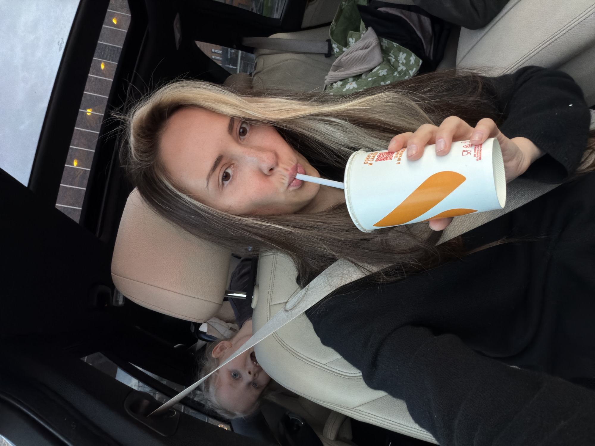 The image captures a casual moment inside a car, featuring a woman in the foreground and a child in the background.
The main subject is a young woman, positioned at an angle, looking directly at the camera with a playful expression, her lips puckered around a white straw. She has long, straight hair with prominent blonde highlights against a darker brown base. She wears a black long-sleeved top. Her right hand, with natural nails, holds a white and orange cup, from which she is drinking.
In the background, behind the woman and slightly to her right, a young child with blonde hair and wide blue eyes is visible, peeking from behind a light-colored car seat. The child's expression is one of curiosity or surprise. A white seatbelt diagonally crosses the foreground, partially obscuring the child and the car seat.
The setting is the interior of a car, characterized by light-colored leather seats and dark trim. Through what appears to be a sunroof or panoramic roof on the left side of the frame, a cloudy sky is visible, along with a pattern of bright yellow lights, possibly reflections or decorative elements on the roof's shade. On the back seat, behind the woman, some folded items are visible, including a green and white floral patterned fabric.
The cup the woman is holding has red Cyrillic text visible on its side. Close inspection reveals phrases such as "БОНУСЫ И СКИДКИ" (Bonuses and Discounts), "Сканируй QR" (Scan QR), and "ТОВАР В ПРИЛОЖЕНИИ" (Product in the Application), indicating a loyalty program or offer. This text, combined with the given location context, suggests the image was taken in Moscow, Russia. The time of day appears to be daytime, under overcast or cloudy weather conditions.
