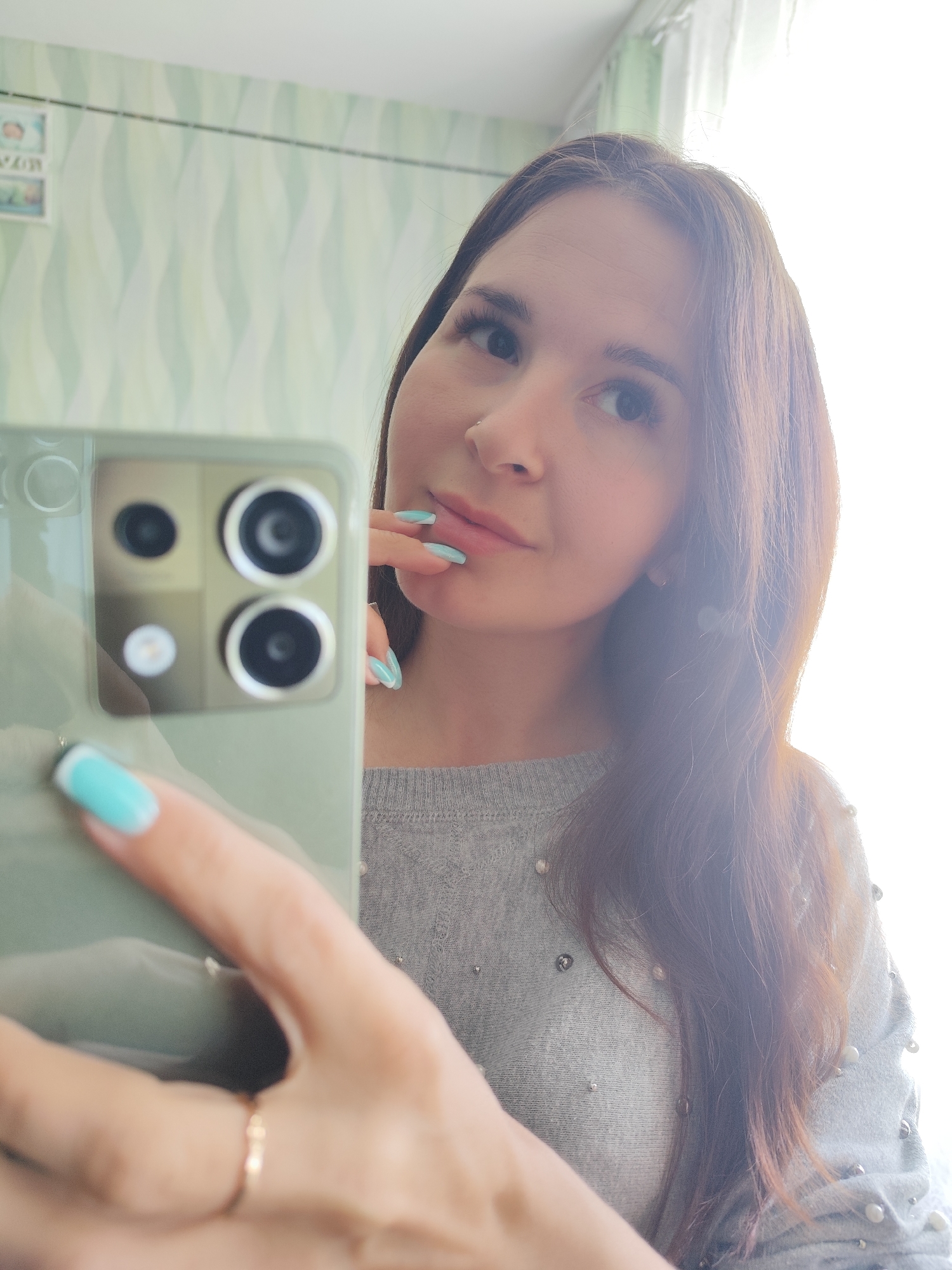 A woman is taking a selfie with a smartphone. The phone is held up in the left foreground, and the camera array is visible on the back. The woman is wearing a gray sweater with small, possibly pearl-like, embellishments. Her hair is long and brown, and she has light blue nail polish. The background is a light green wall with a patterned design, and a window is visible on the right. The location cannot be determined as Фоки, Russia based on the image content.